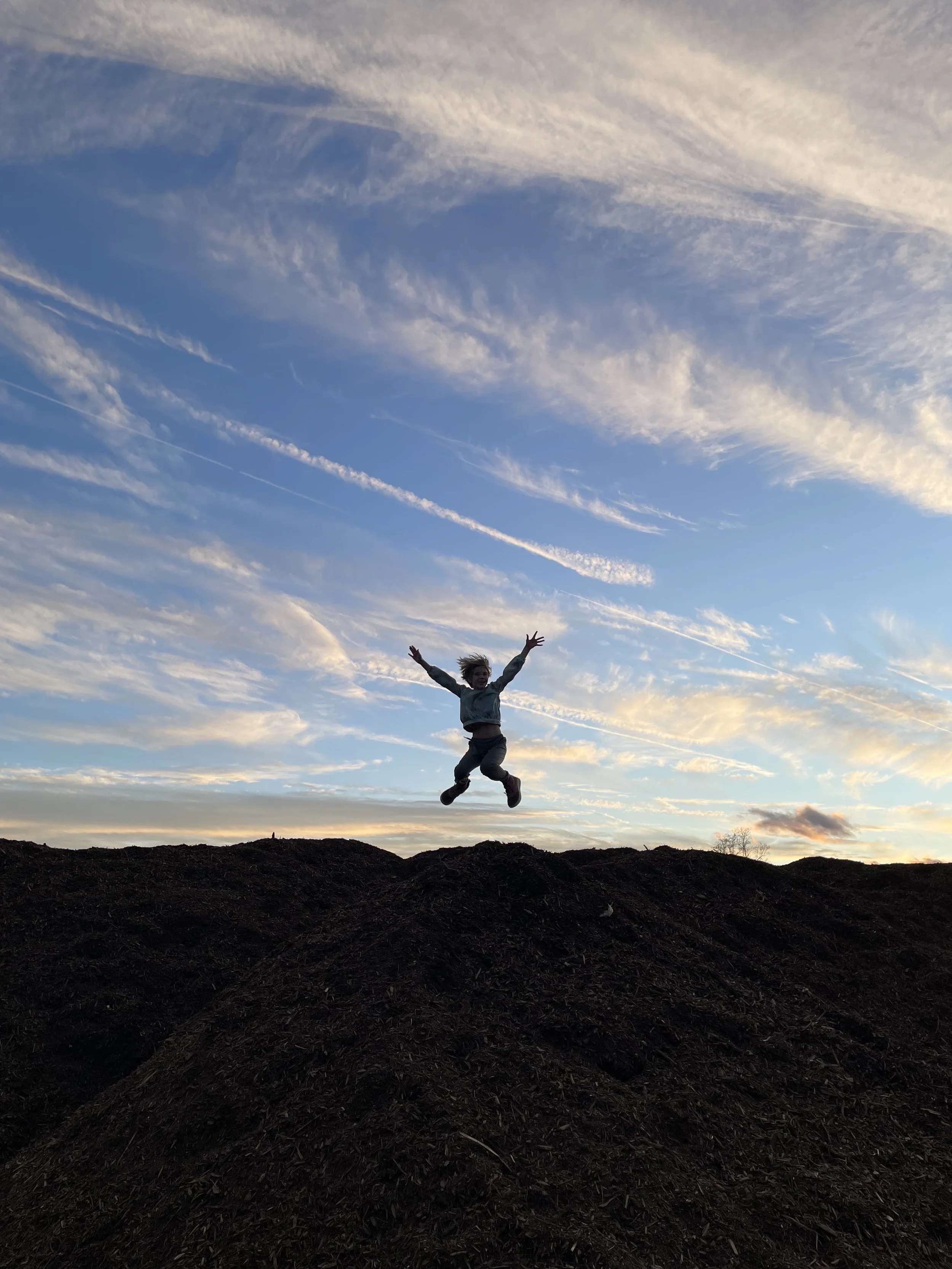 A person jumping in the air on a mound of dirt, with the sky and clouds at sunset in the background.