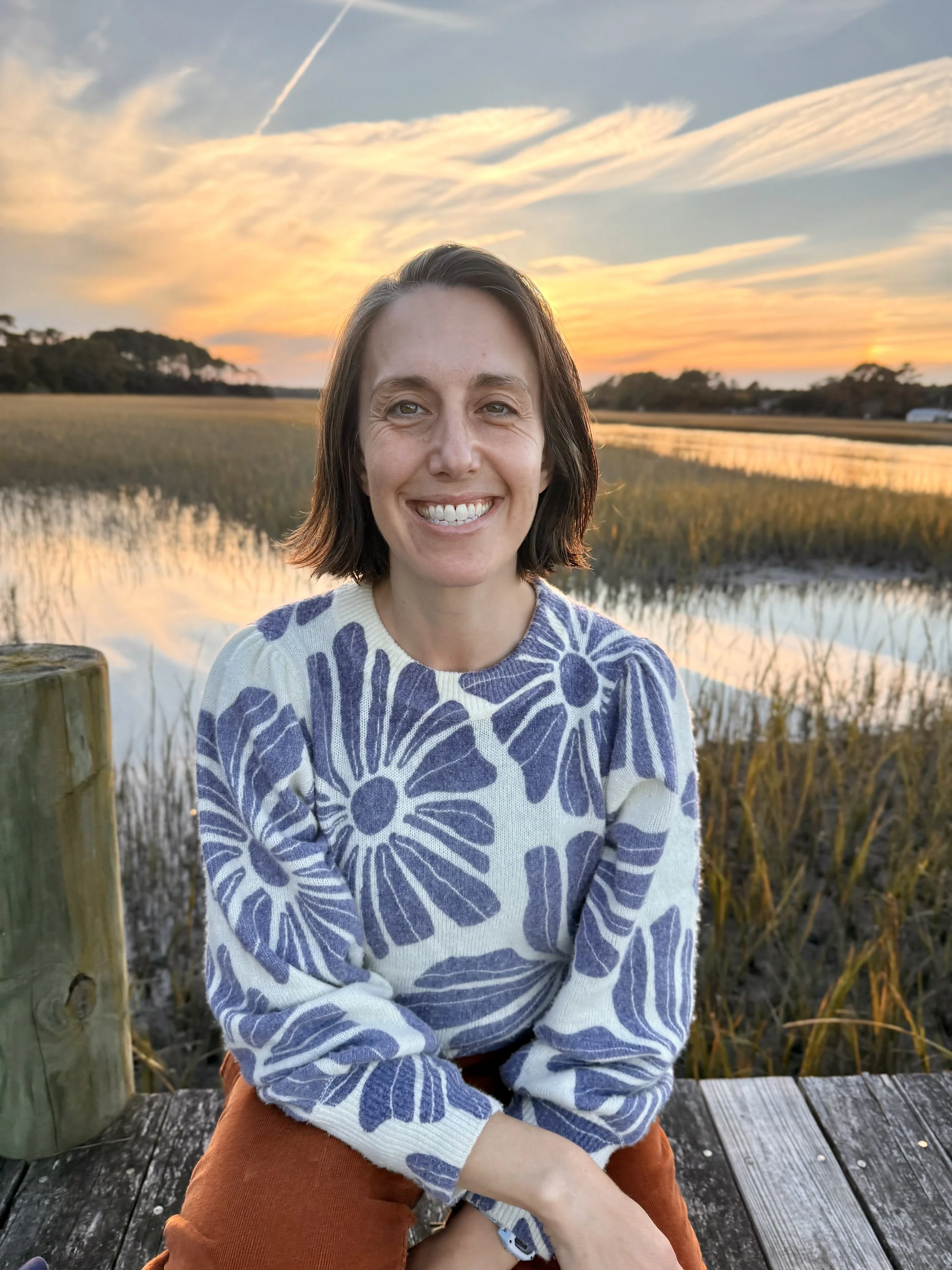 A woman with shoulder-length brown hair smiling at the camera, sitting on a wooden dock at sunset with marshlands and a colorful sky in the background.