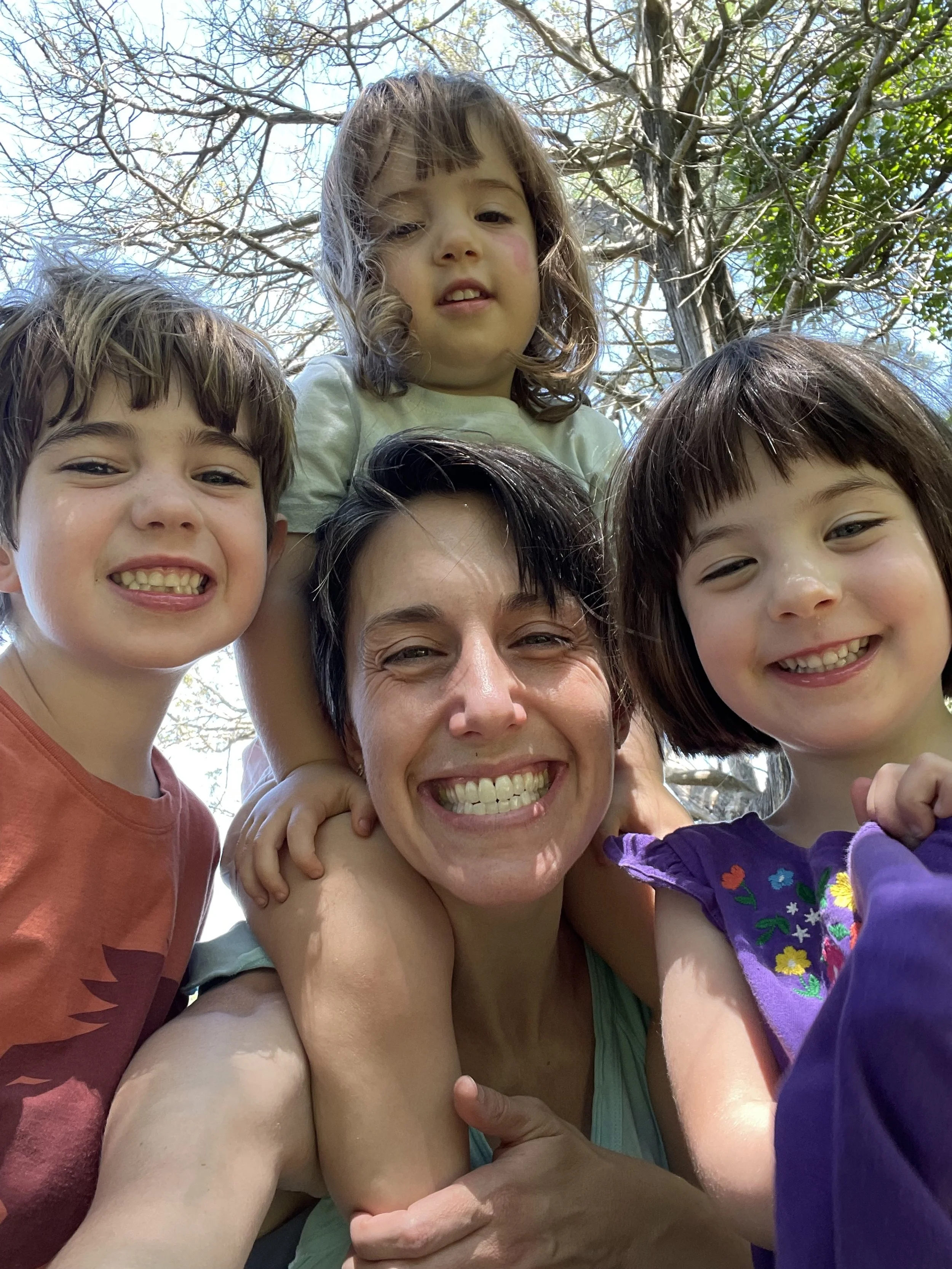 A group of five smiling people, four children and one adult woman, taking a selfie outdoors with trees and blue sky in the background.