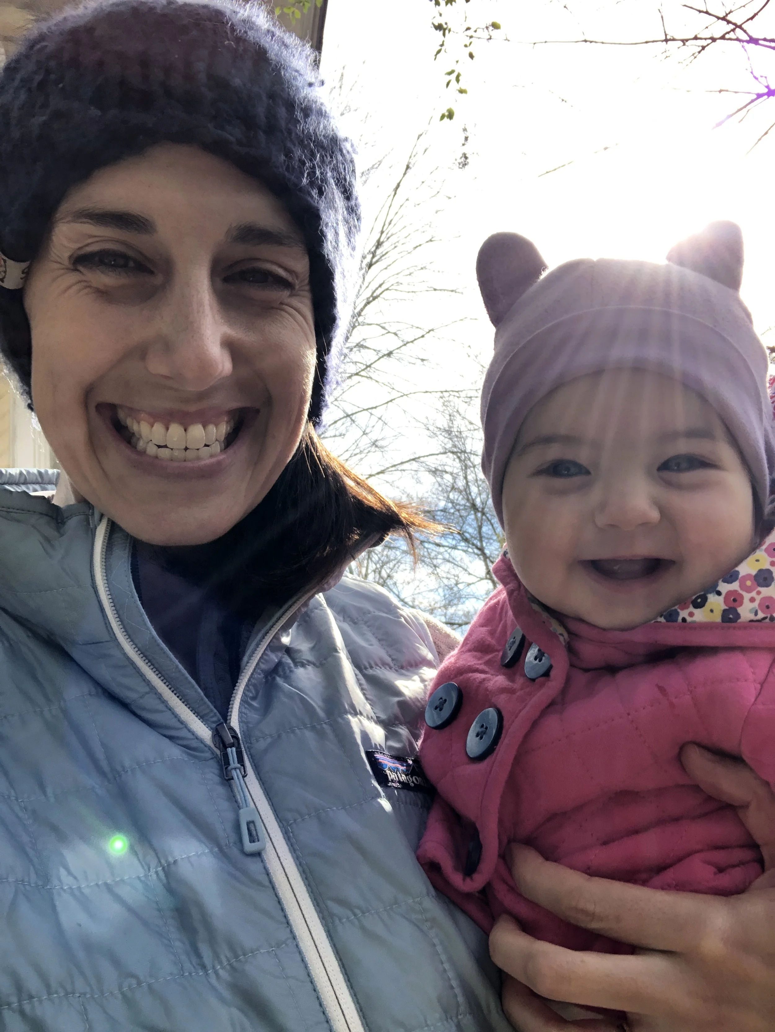 A woman smiling and holding a young girl outdoors in front of a bright sky and leafless trees.