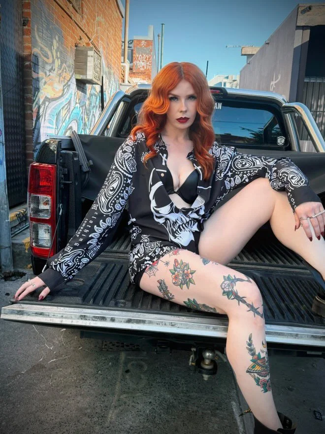 red-haired model siting in boot of black truck, wearing long-sleeved collar black and white bandit shirt with white skull and swirly pattern inspired by Los Angeles street gang bandannas