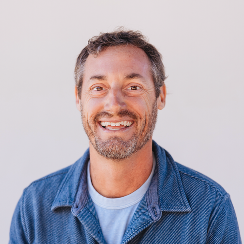 Smiling man with short gray hair and beard wearing a denim jacket and a light-colored shirt.