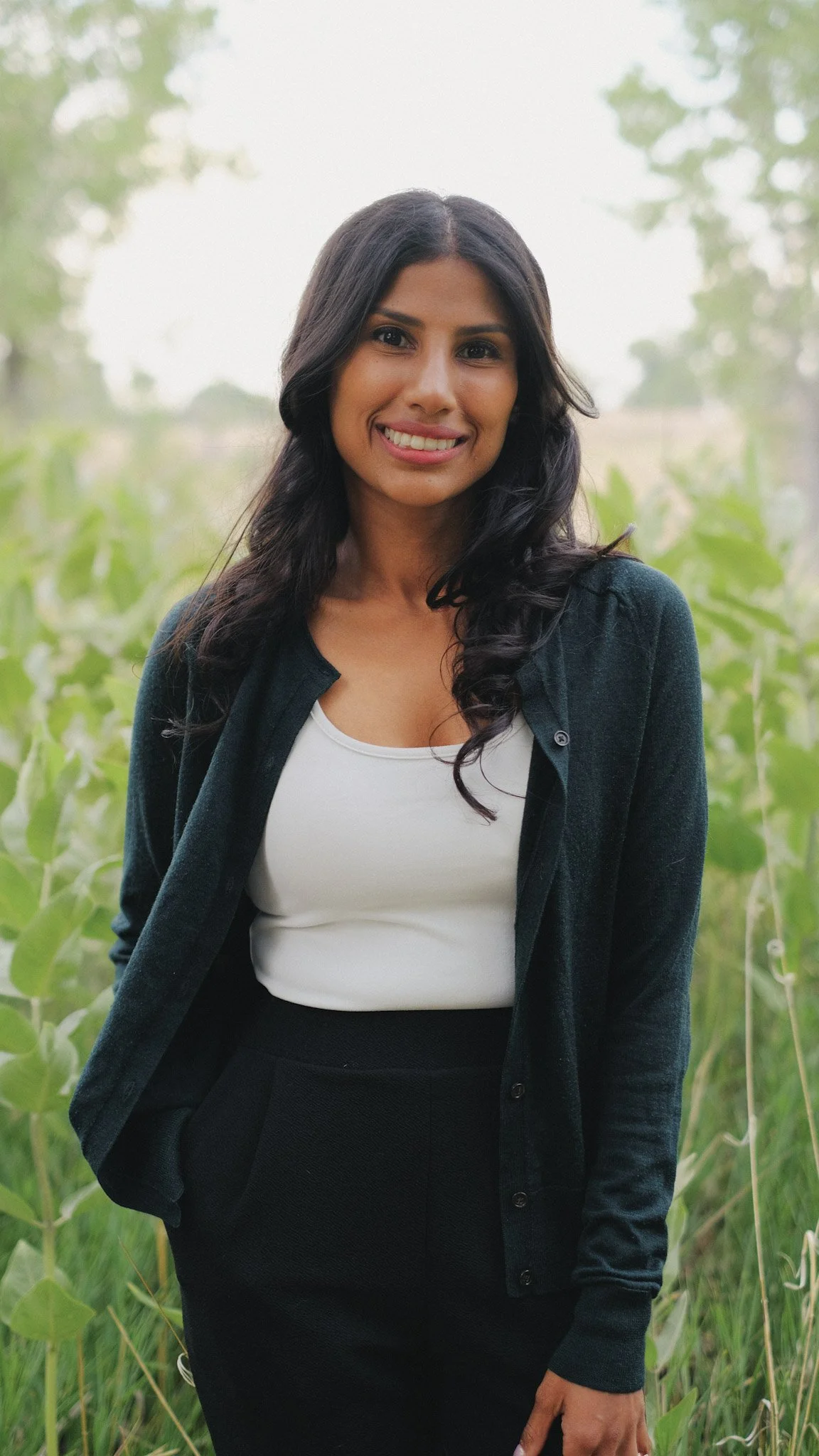 A woman with long dark hair wearing a white top, black cardigan, and black skirt standing outdoors in a field with tall green plants and trees in the background.