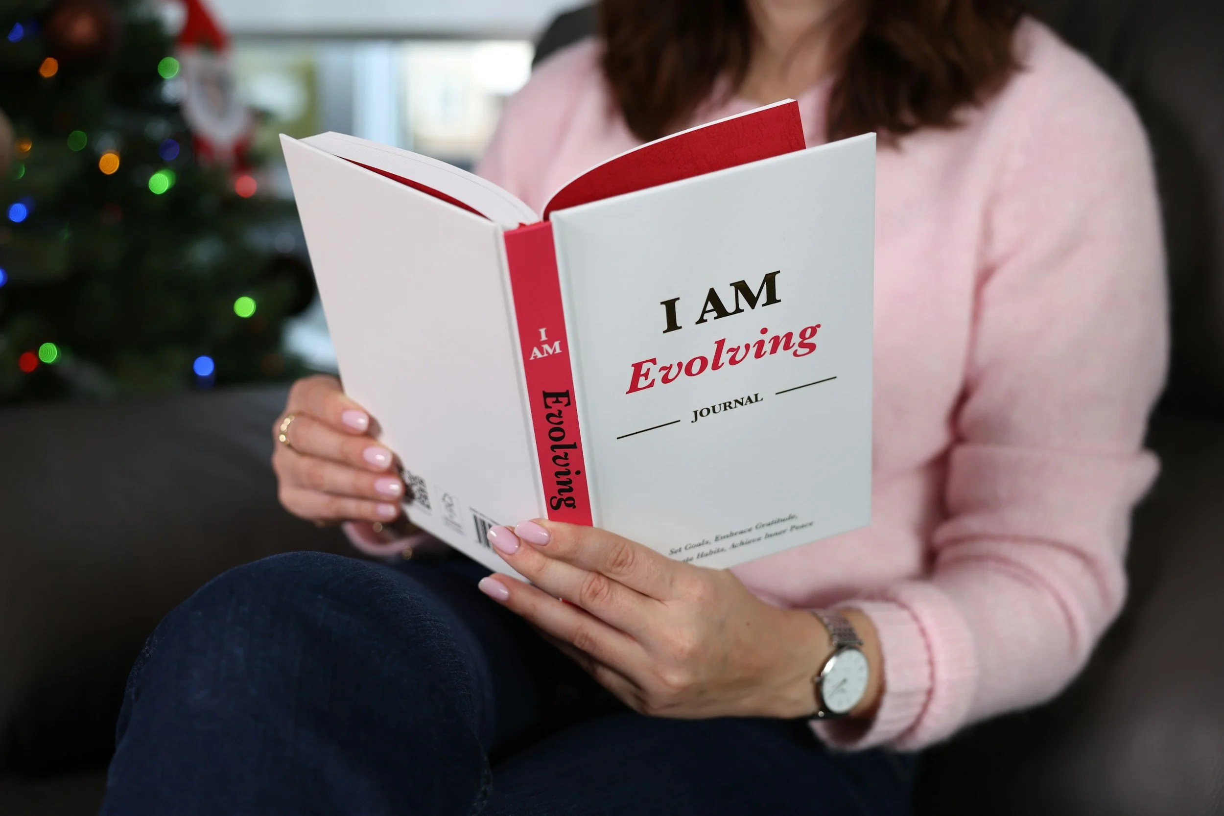 Person in pink sweater reading a book titled 'I AM Evolving' with a Christmas tree in the background.