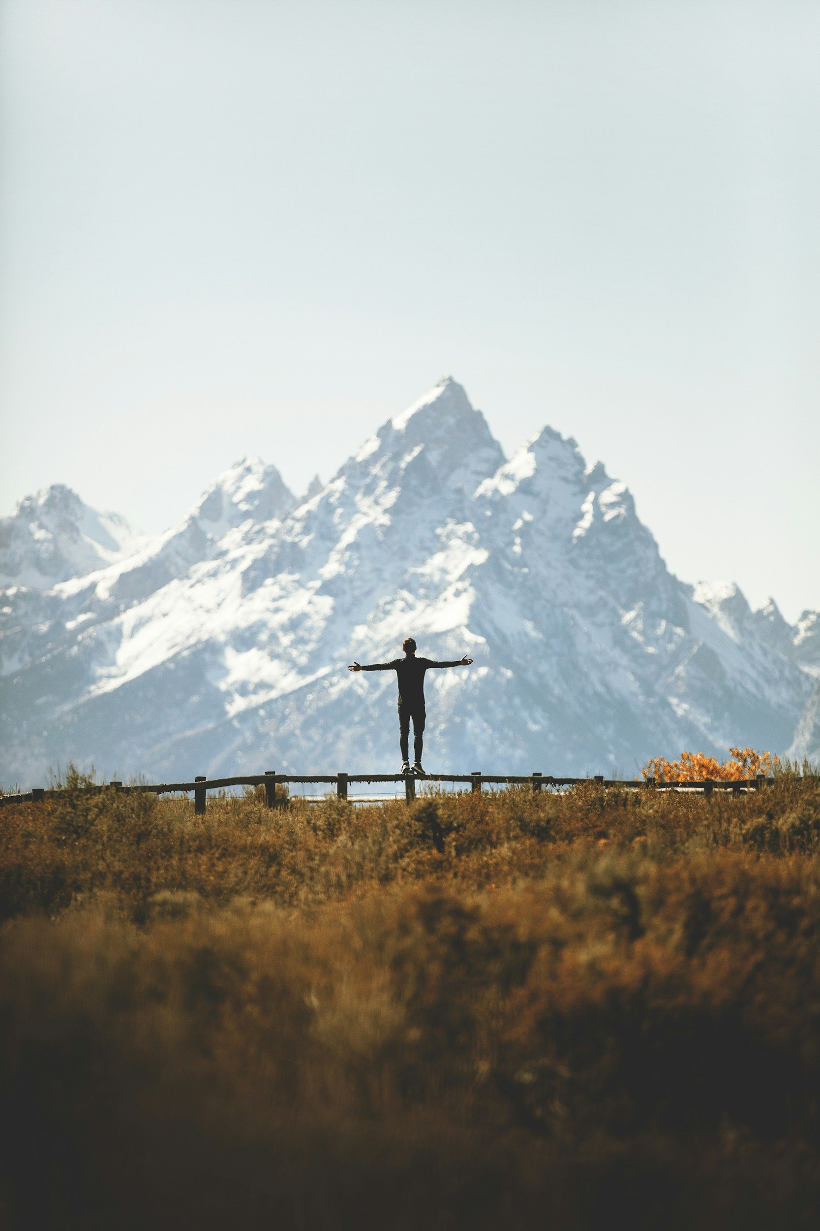 Person standing with arms outstretched on a wooden fence in front of snow-capped mountains, clear sky, and autumn foliage.
