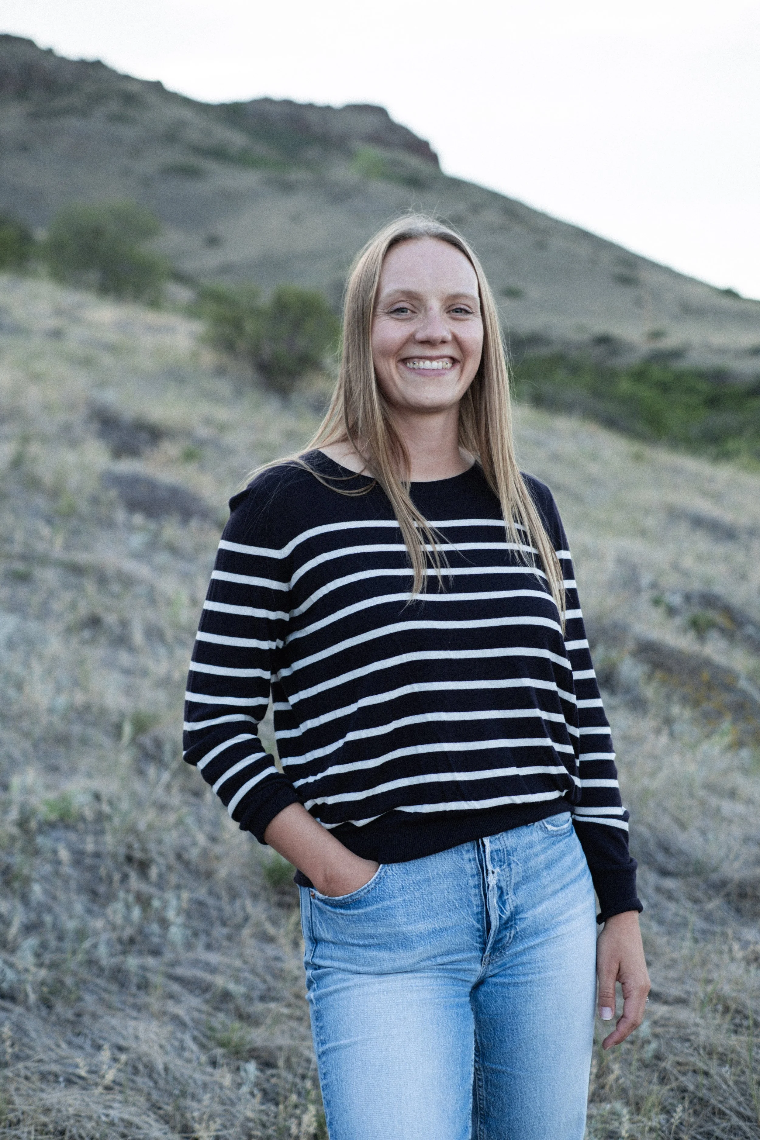A young woman with long blonde hair smiling while standing outdoors on a grassy hillside with sparse vegetation and hills in the background.