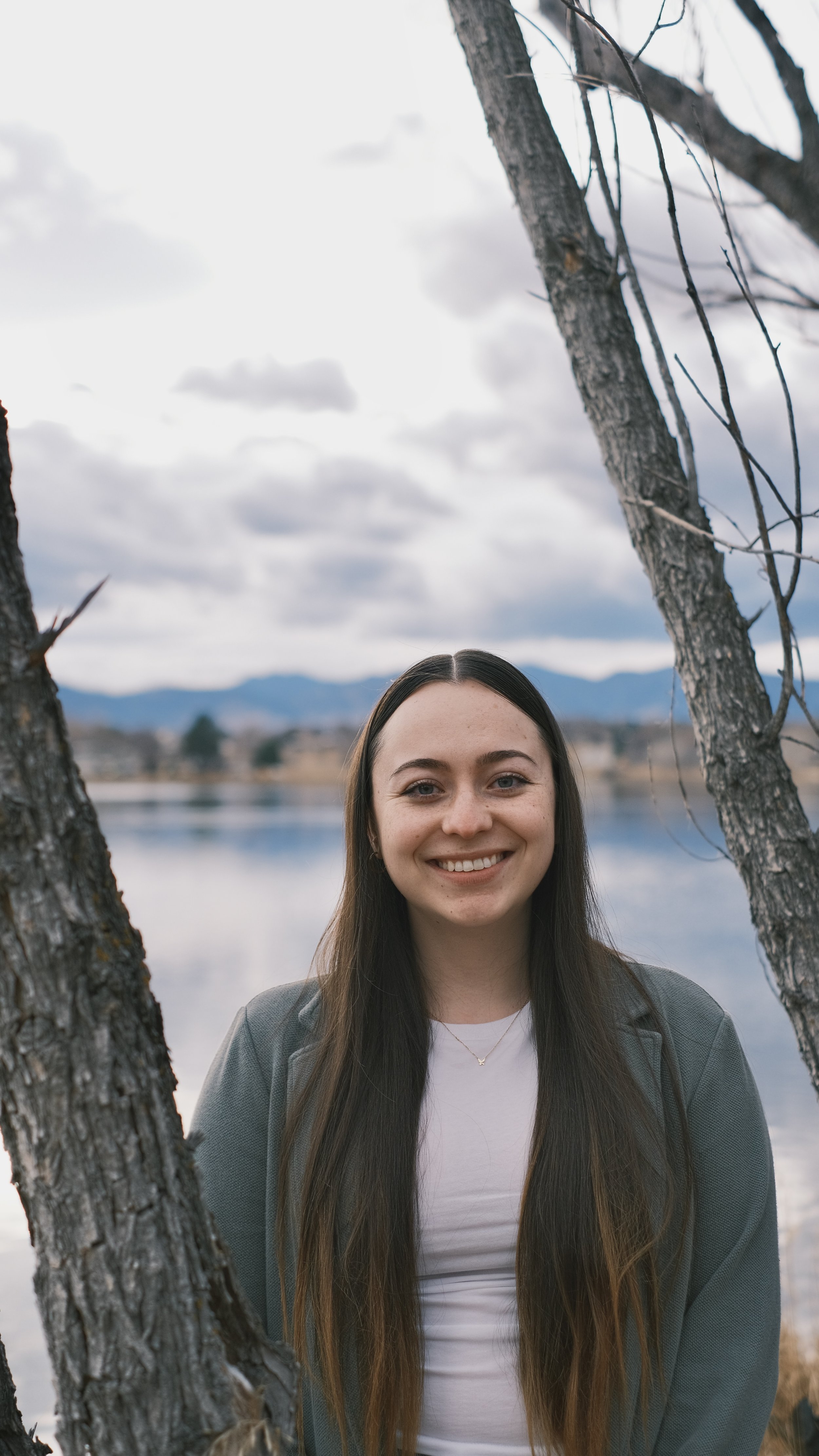 A young woman with long dark hair smiling outdoors by a lake with mountains in the background, framed by two trees.