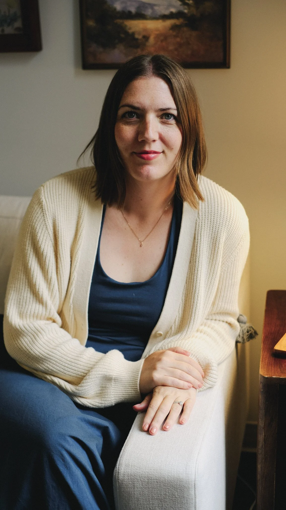 A woman with shoulder-length brown hair, wearing a beige cardigan and a navy top, sitting on a white chair in a room with paintings on the wall. She is looking at the camera, with hands resting on her lap, wearing a ring and a necklace.