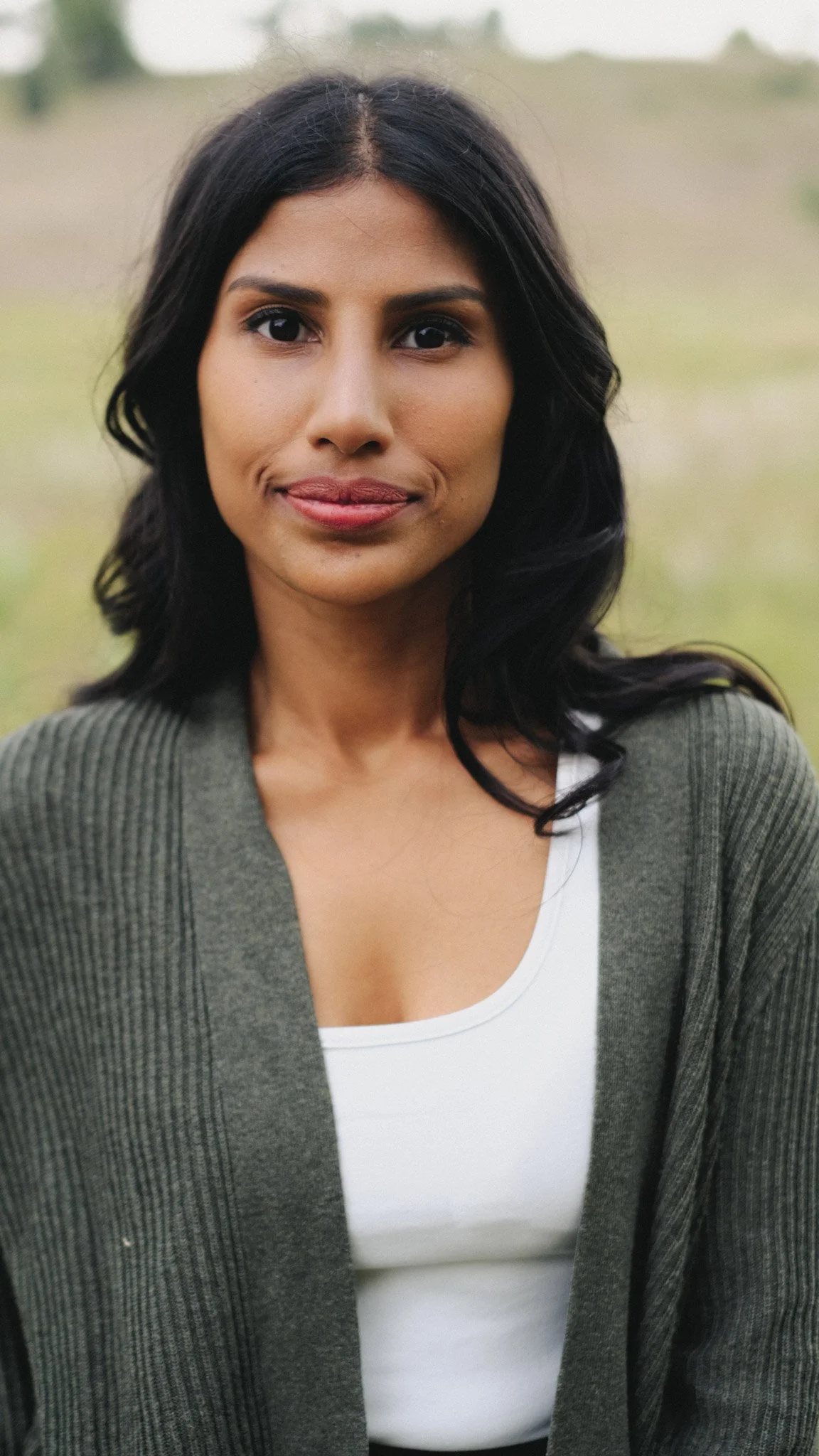 A young woman with dark hair and brown skin standing outdoors, wearing a white top and a dark green cardigan, with a blurred background of grass and trees.