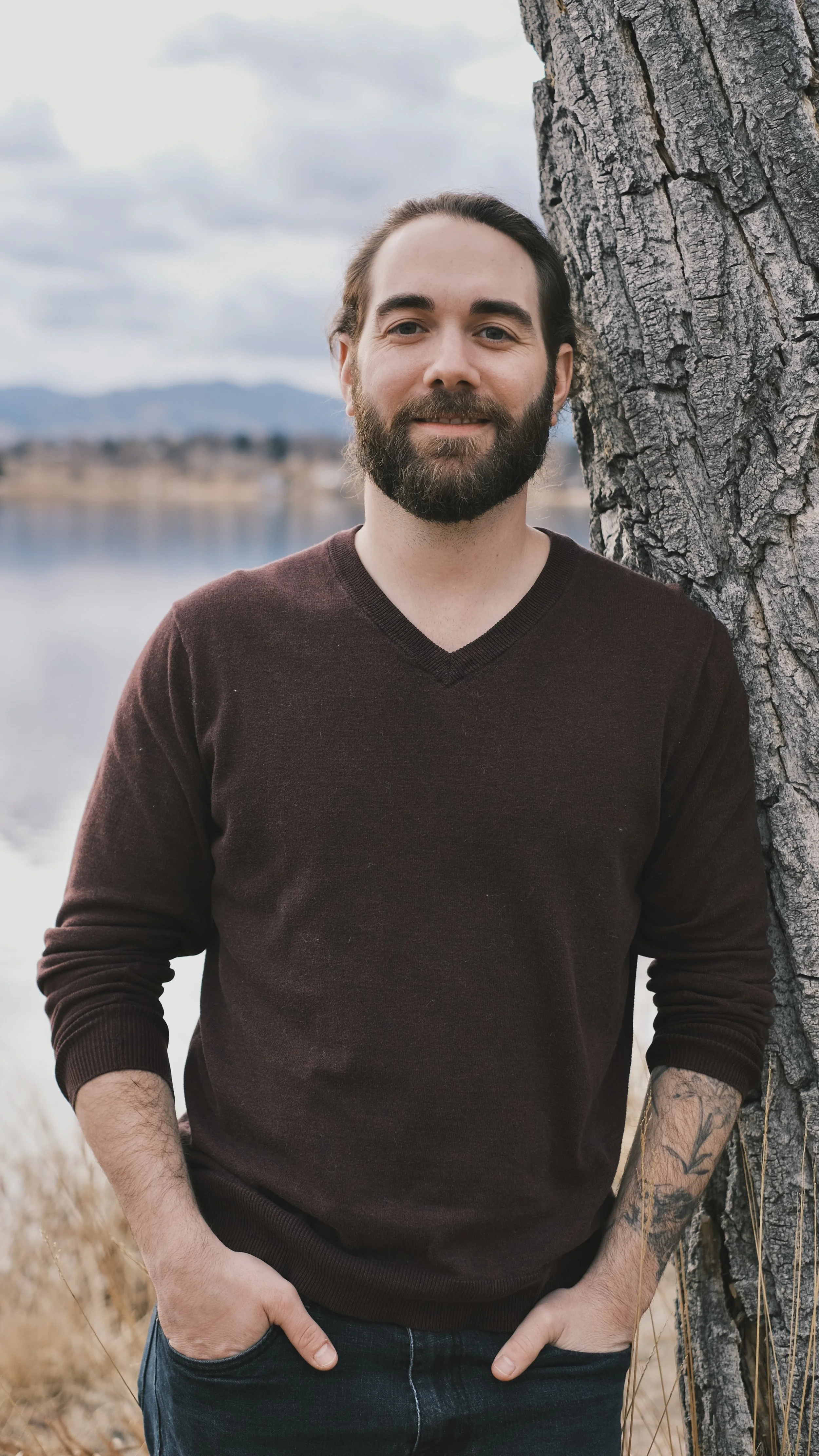 A man with a beard and long hair standing outdoors near a tree by a lake, with mountains and cloudy sky in the background.