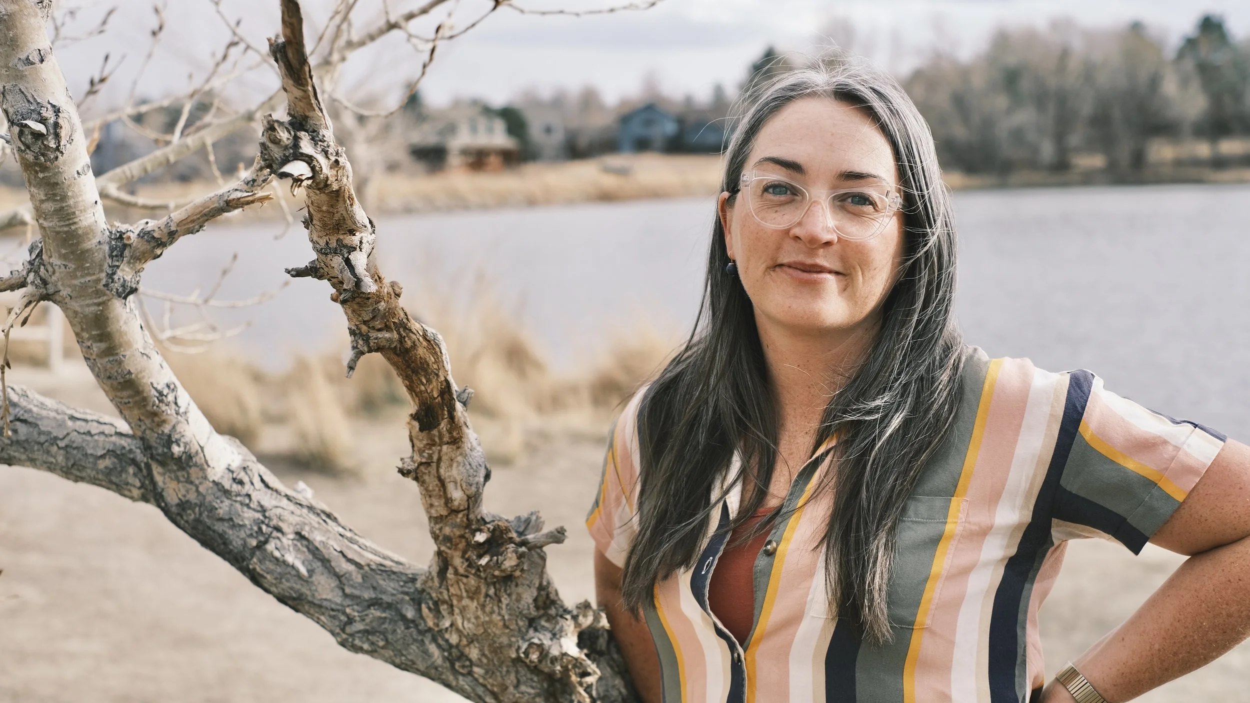 Professional Headshot Example: Woman with long gray hair and clear glasses smiling outdoors near a lake with leafless trees and houses in the background.