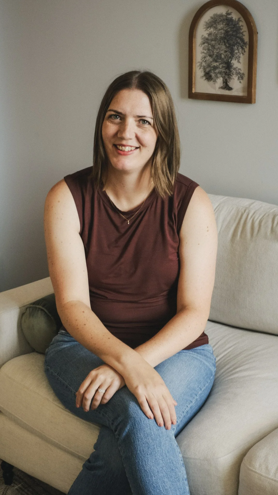 A woman with shoulder-length brown hair, wearing a sleeveless maroon top and blue jeans, sitting on a beige couch with a gray cushion, smiling at the camera. There is a framed tree picture hanging on the white wall behind her.
