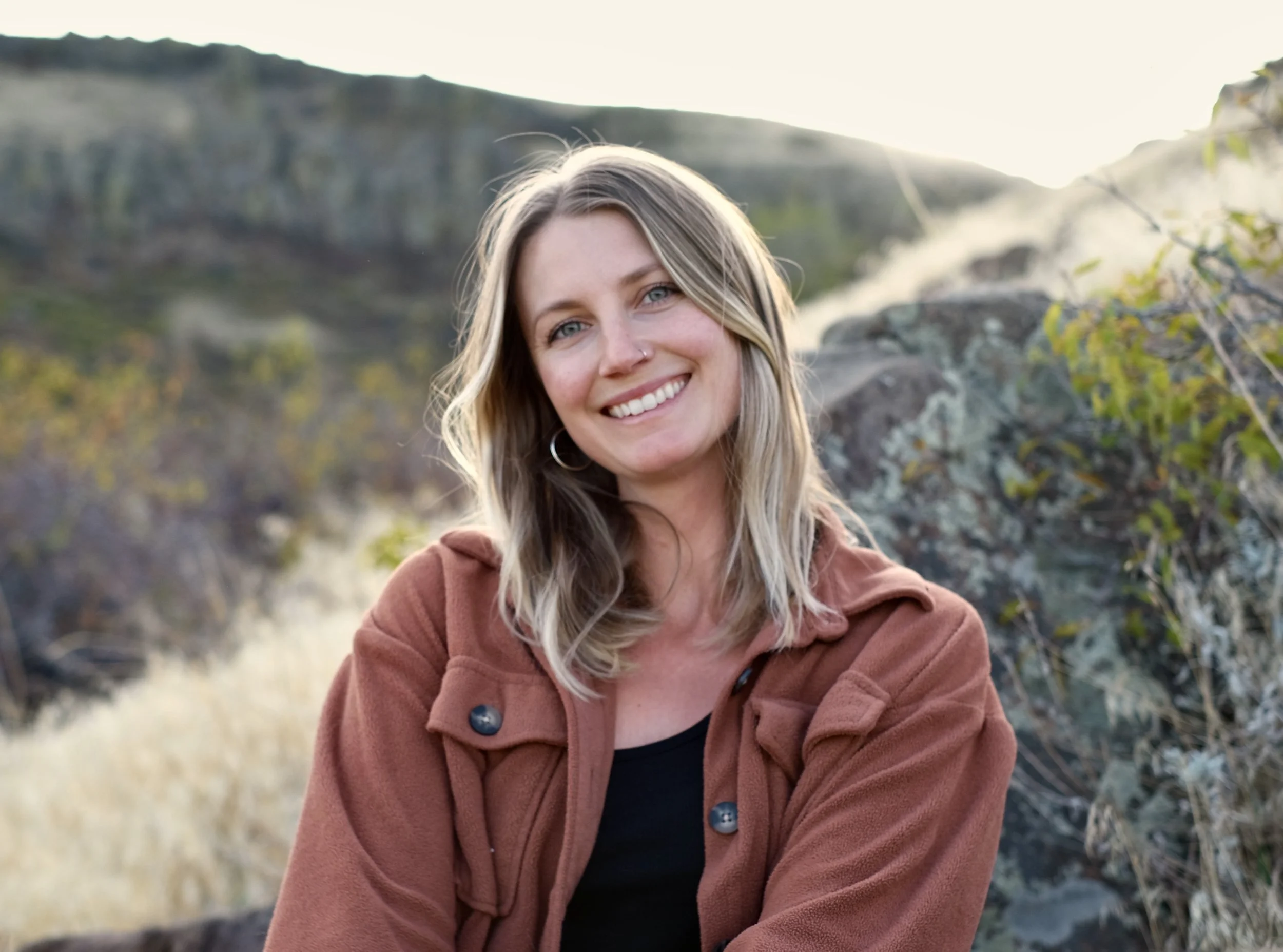 Smiling woman with shoulder-length blonde hair, wearing a brown jacket and black top, outdoors in a natural setting with rocks, trees, and a scenic backdrop.
