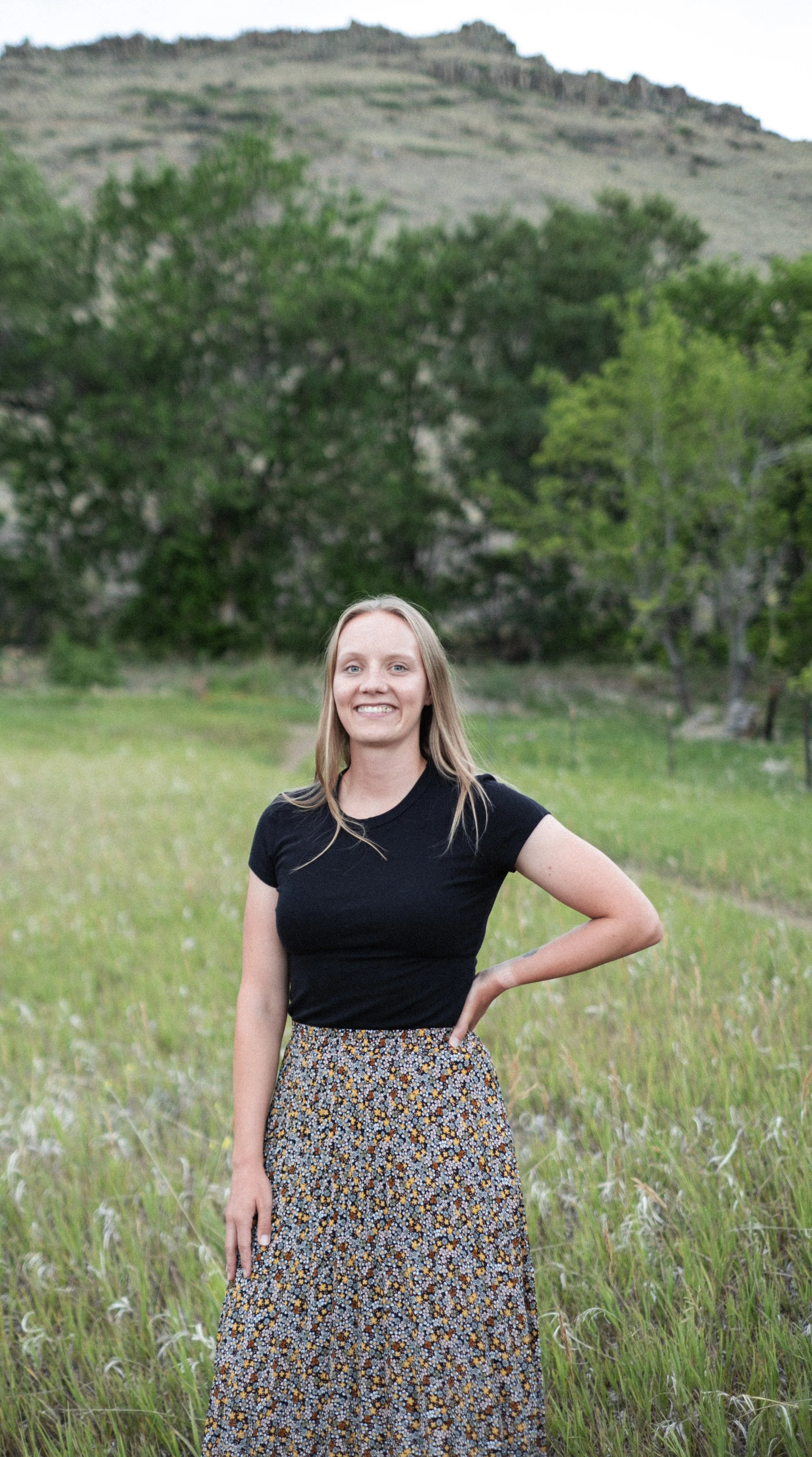 Smiling woman with blonde hair, wearing a black shirt and a long floral skirt, standing in a grassy field with trees and mountains in the background.