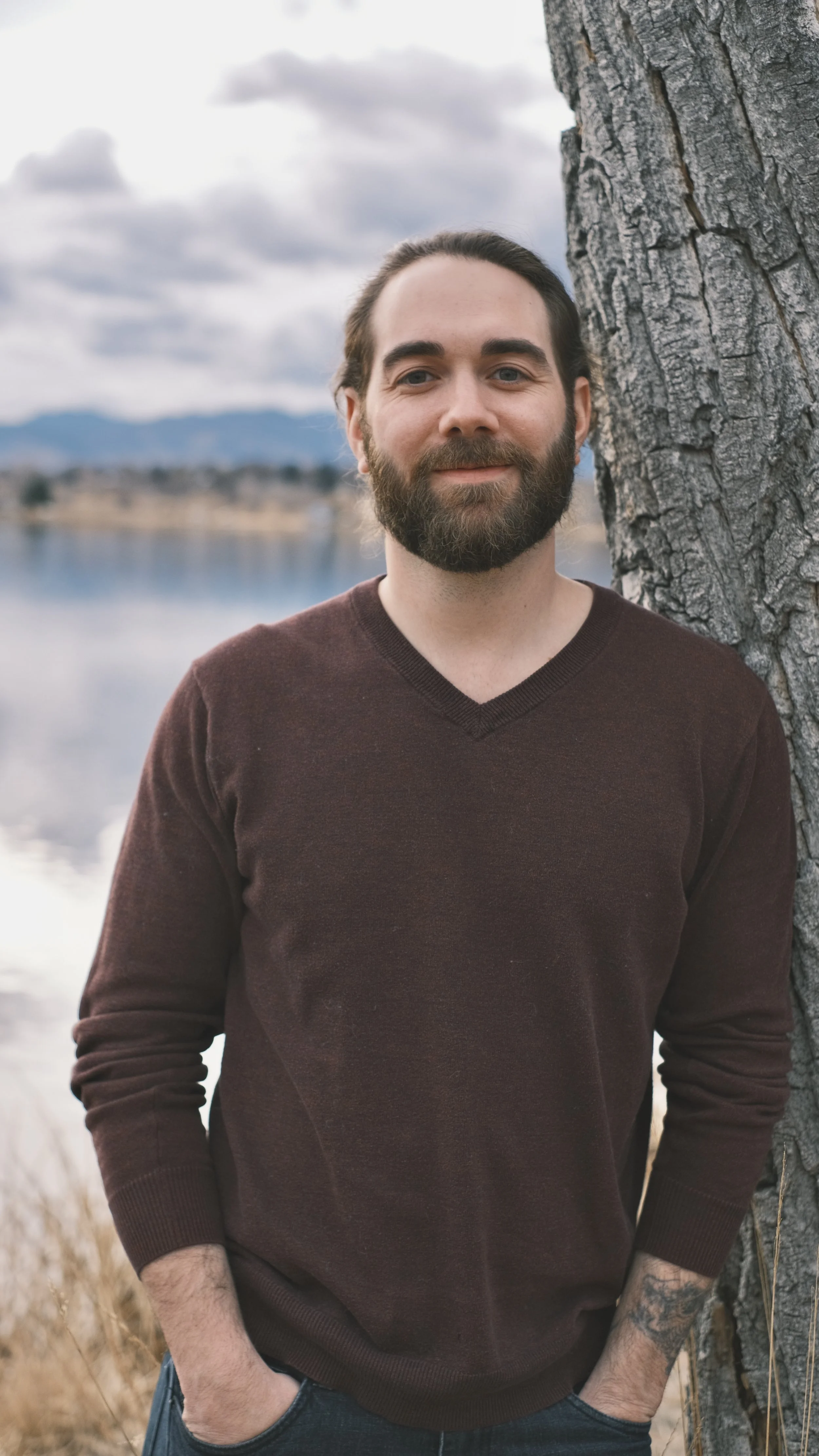 Professional Headshot Example: A man with a beard wearing a brown sweater standing outdoors near a tree with a lake and mountains in the background.