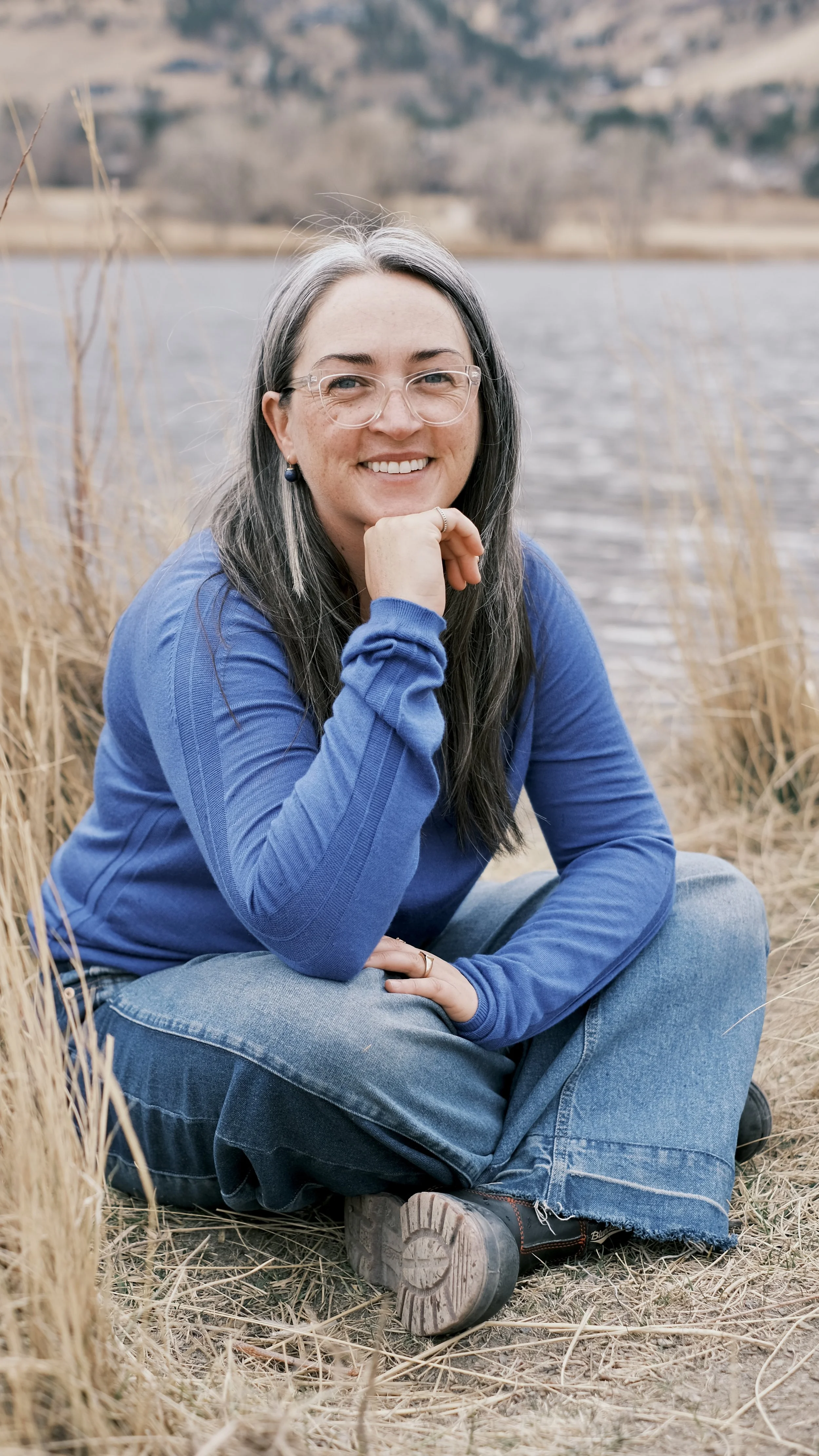 Woman sitting on ground beside a lake, smiling, wearing glasses, blue long-sleeve shirt, jeans, and boots, with dry grass in the foreground and a blurred landscape in the background.