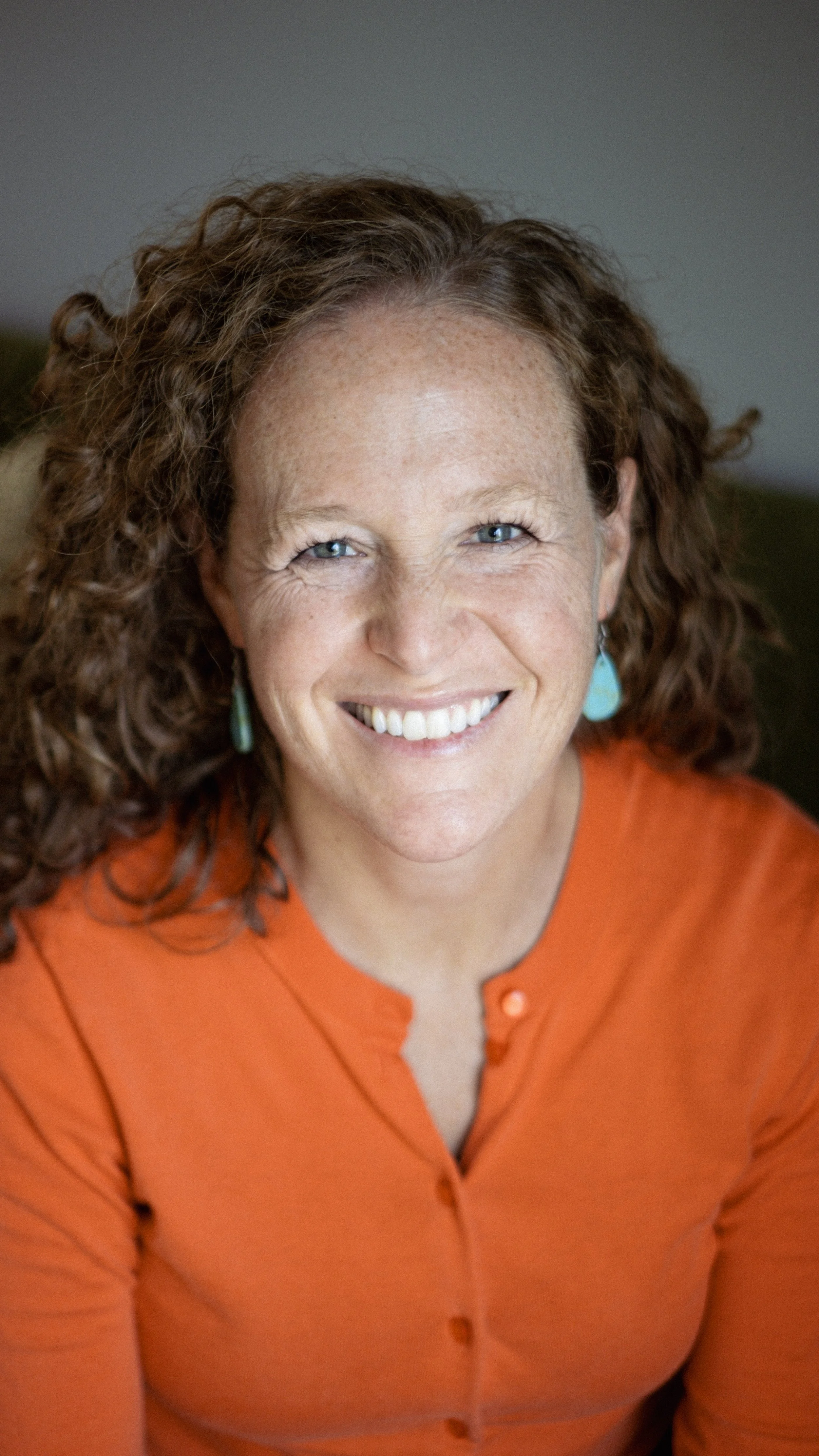 Close-up of a smiling woman with curly red hair, wearing turquoise earrings and an orange shirt.