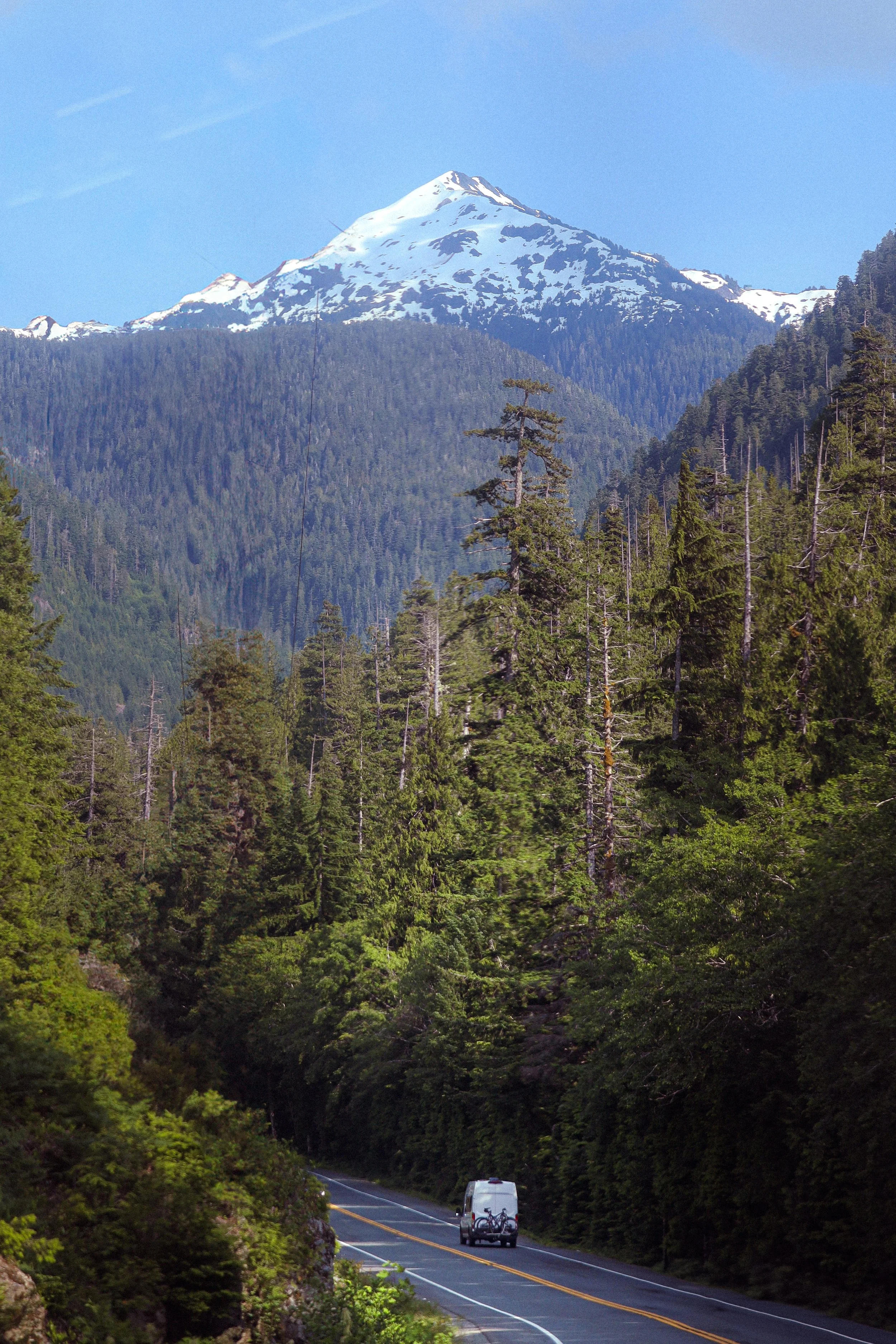 A mountainous landscape with snow-capped peaks, dense green forest, and a winding road with a vehicle traveling on it.