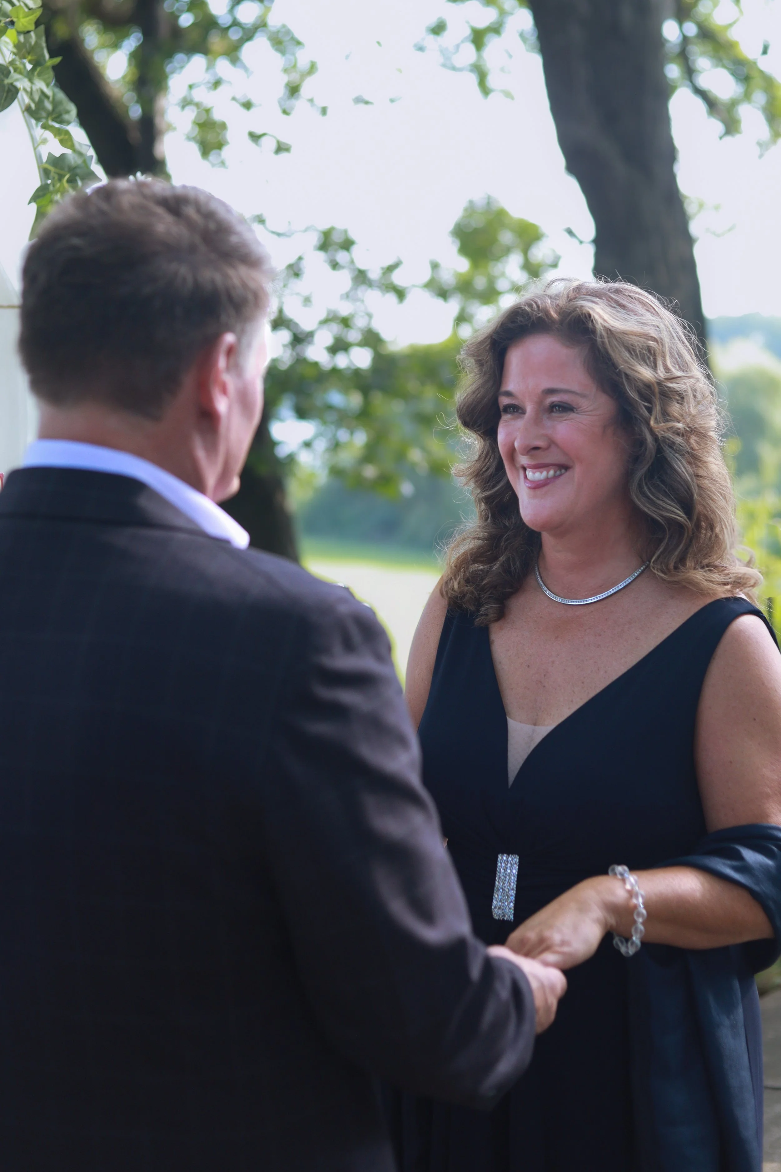 A man and woman exchanging wedding vows outdoors under a tree, with the woman smiling and holding the man's hands.