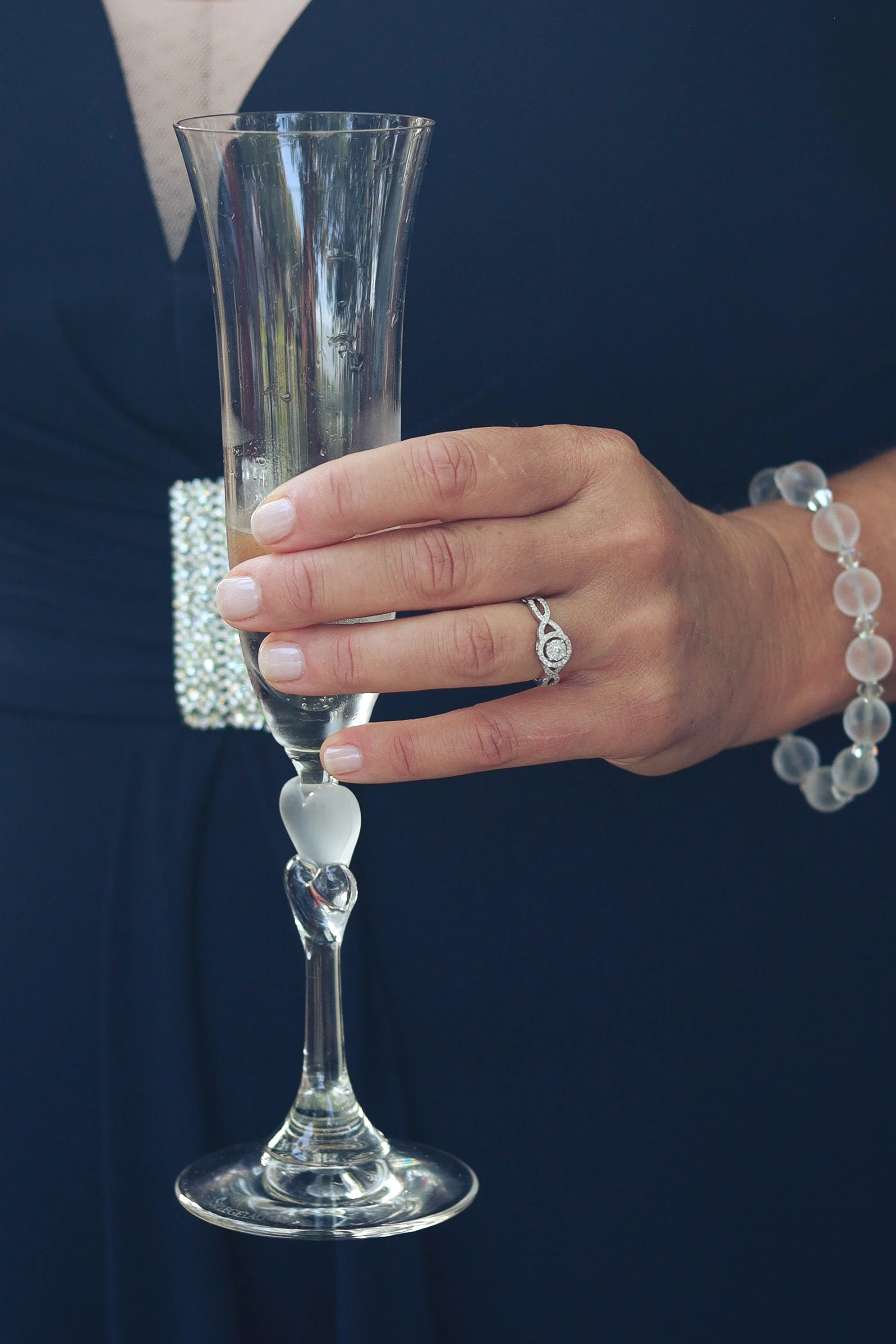 A hand holding a tall, empty champagne flute, wearing a wedding ring and a bracelet, against a dark blue suit with a pocket square.