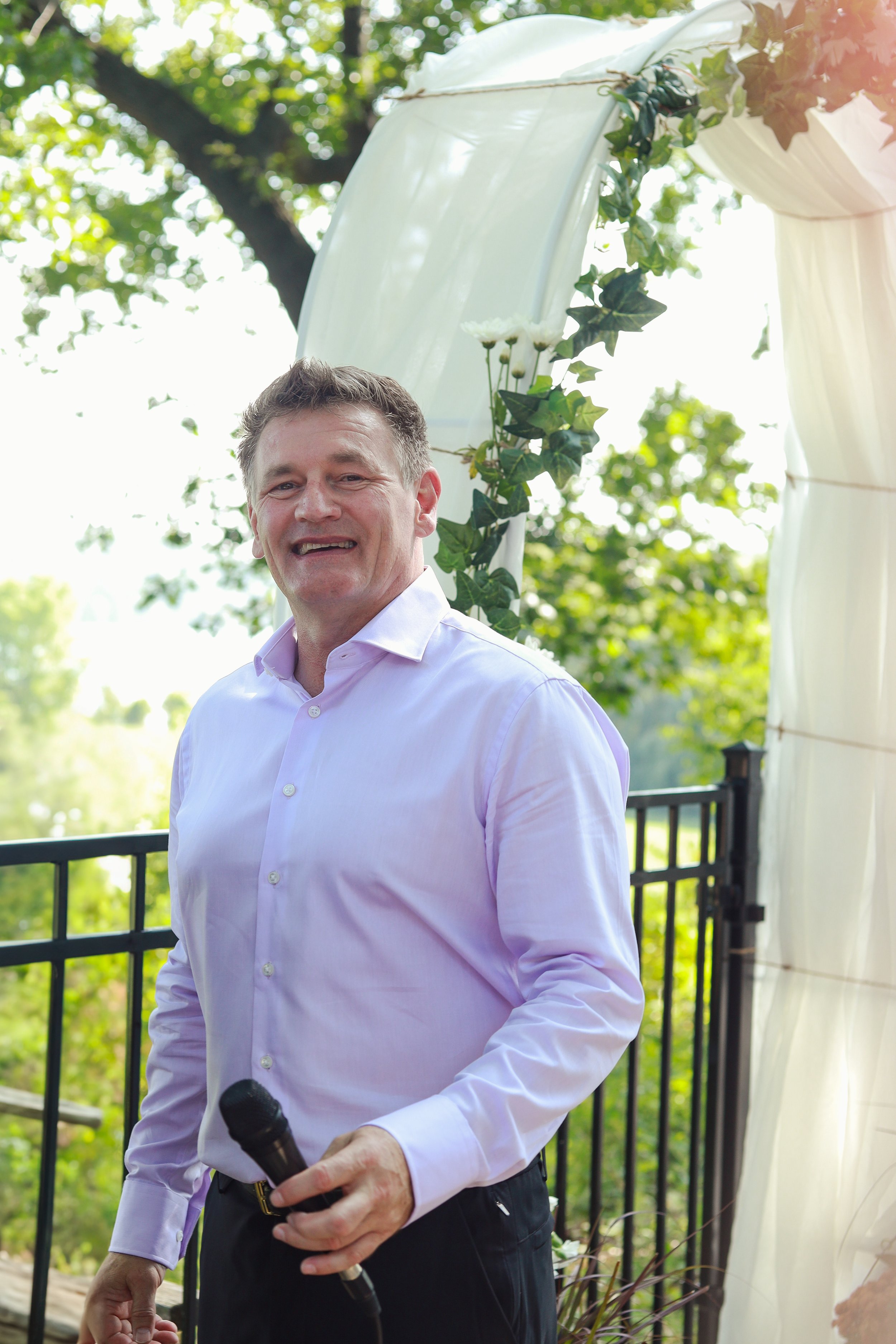 A smiling man in a white shirt holding a microphone outdoors during the daytime at a celebration event. There is a decorated arch with green leaves and white flowers behind him, and some trees with sunlight in the background.