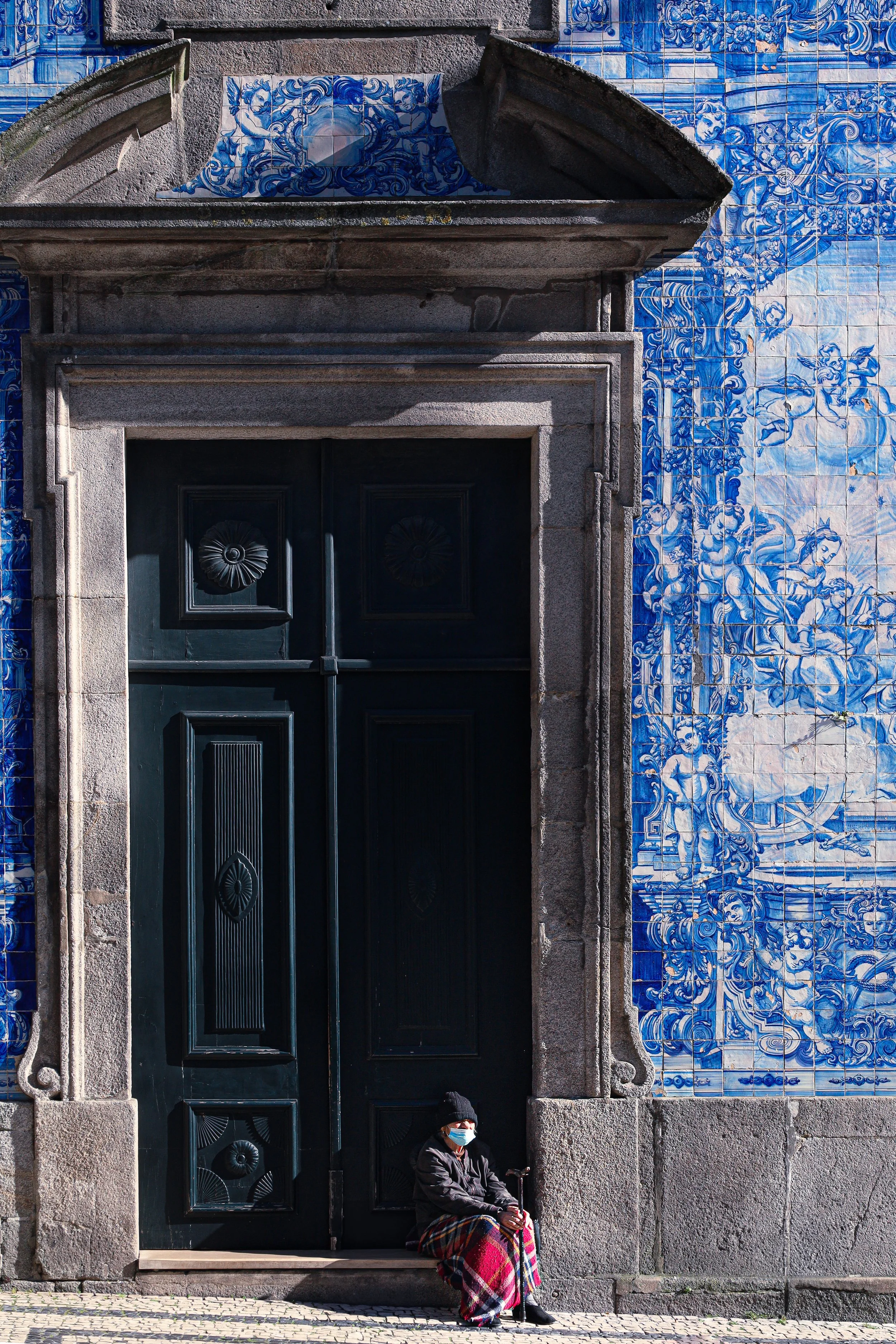 A woman sitting with a cane on stone steps in front of a black door and decorated with blue and white ceramic tiles featuring cherubit and floral designs.