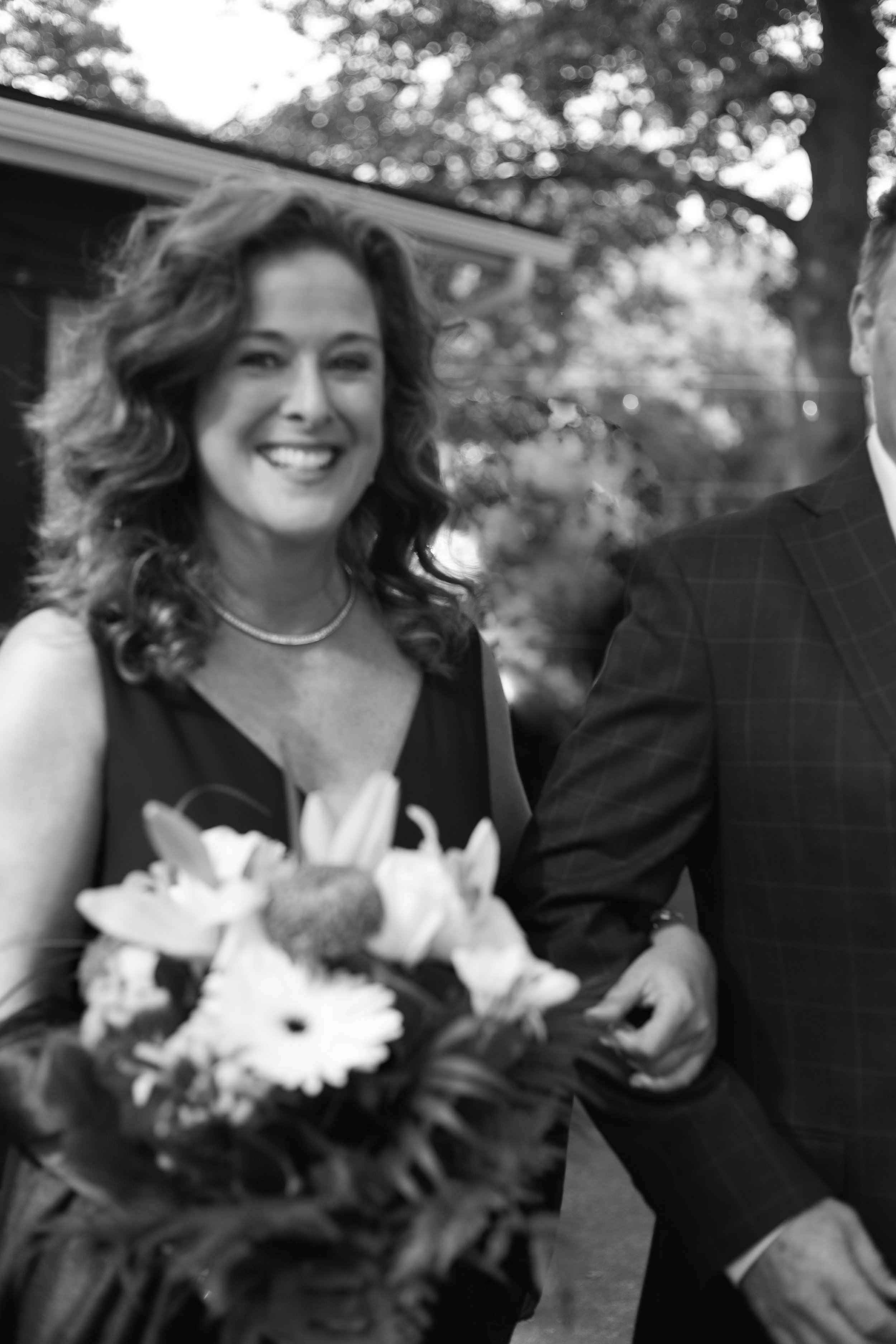 A smiling woman wearing a sleeveless dress holding a bouquet of flowers, standing beside a man in a suit, outdoors with trees in the background.