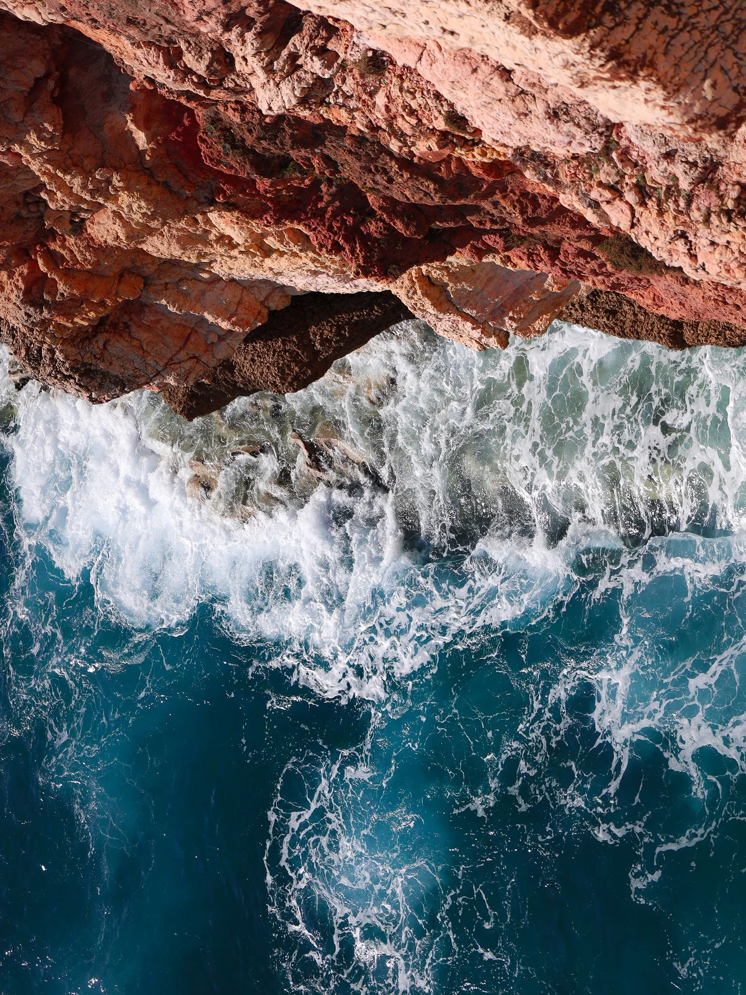 Red rocky cliffs with foamy blue ocean waves crashing below.