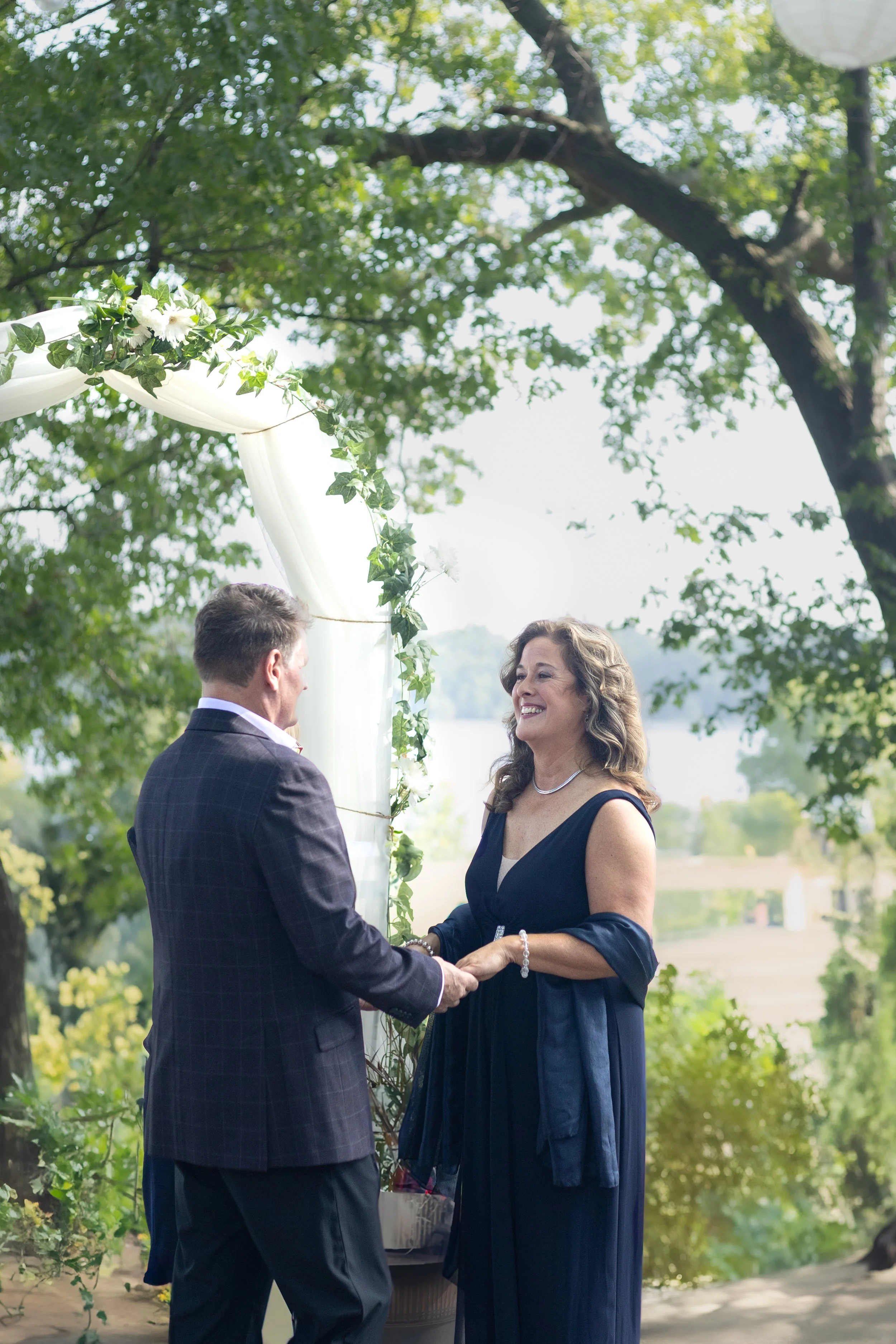 A couple getting married outdoors, holding hands and exchanging vows under a decorated arch with greenery and white flowers, with a river visible in the background, on a sunny day.
