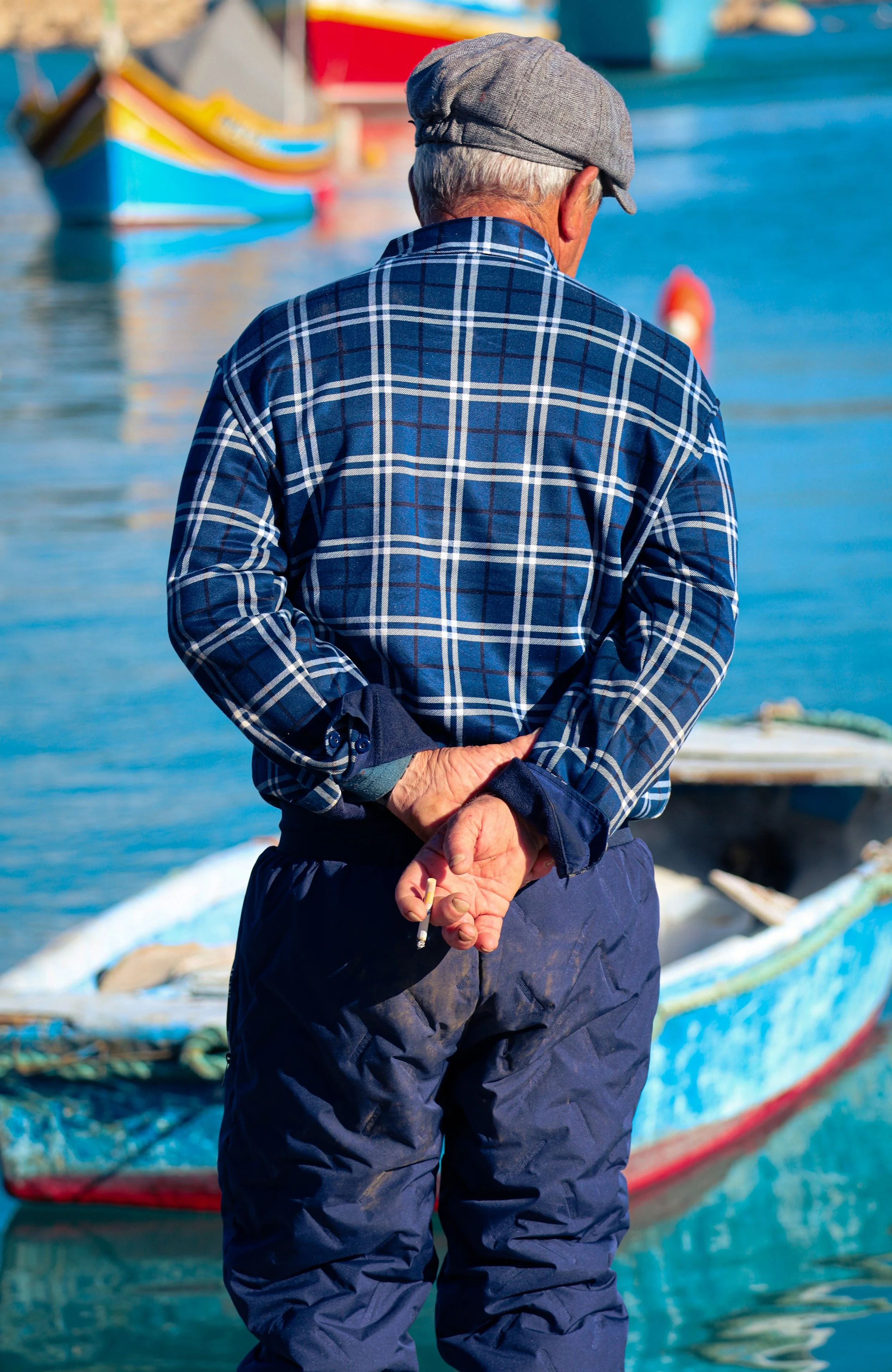 An older man with gray hair wearing a gray cap and a blue plaid shirt stands with his back facing the camera by a harbor. He is holding a cigarette in his right hand behind his back, and there are colorful boats docked in the water behind him.