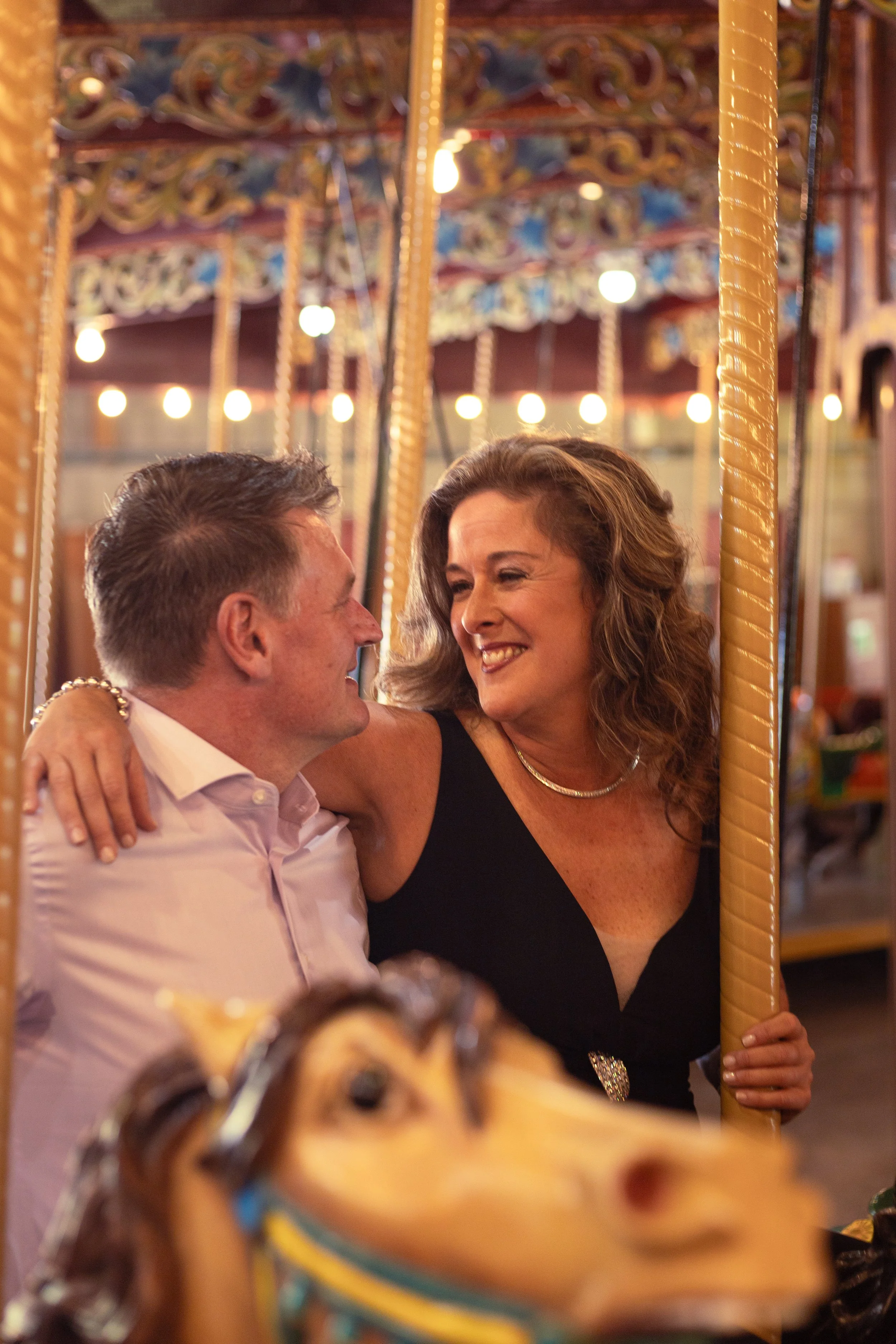 A smiling woman and a man are sitting close together on a carousel ride at night, with the woman holding a carousel pole.