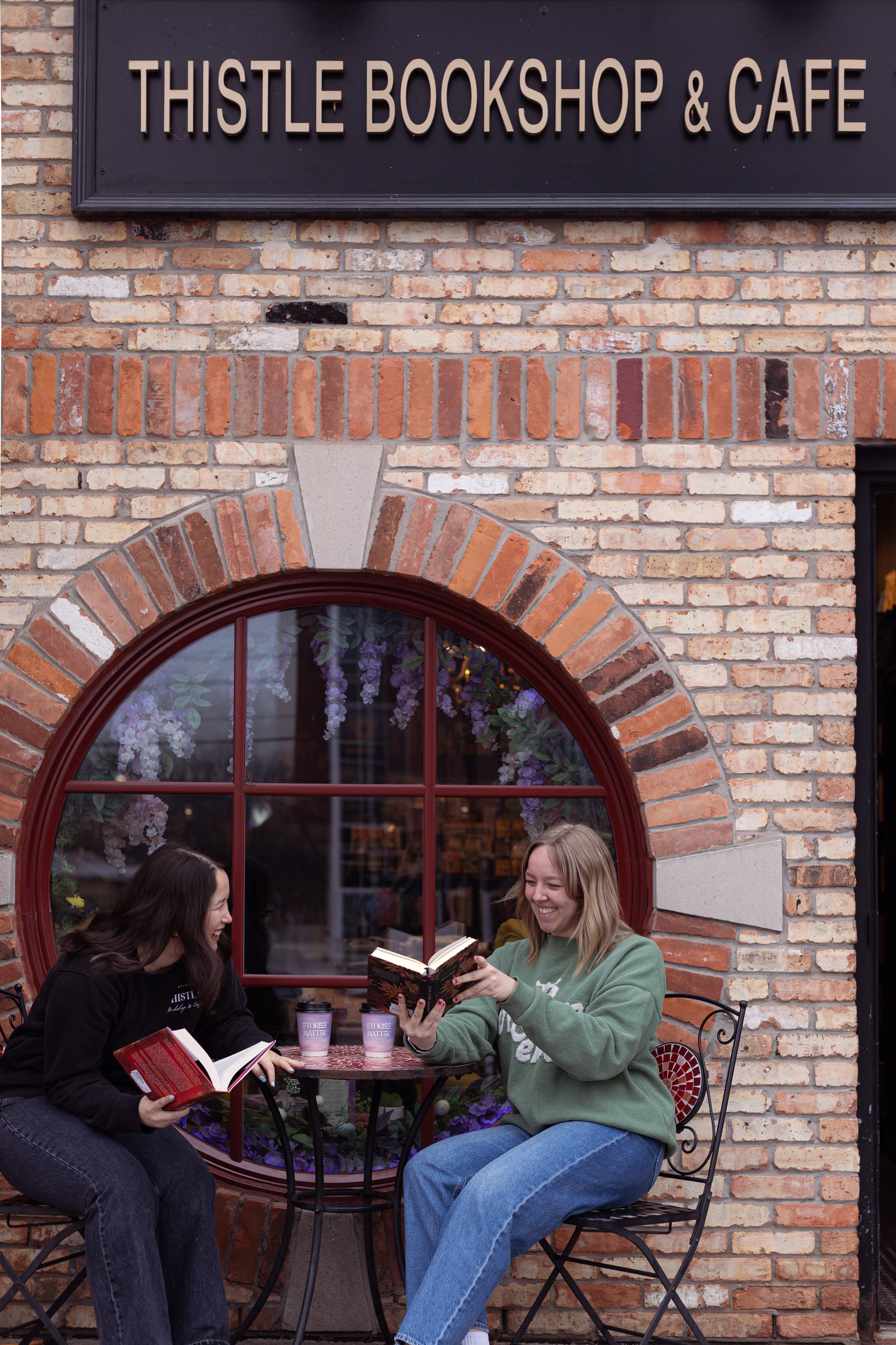 Two women sitting outside at a small round table in front of a brick building, smiling and reading books, with iced beverages labeled "Stokes Matter" on the table, in front of The Thistle Bookshop & Cafe sign.