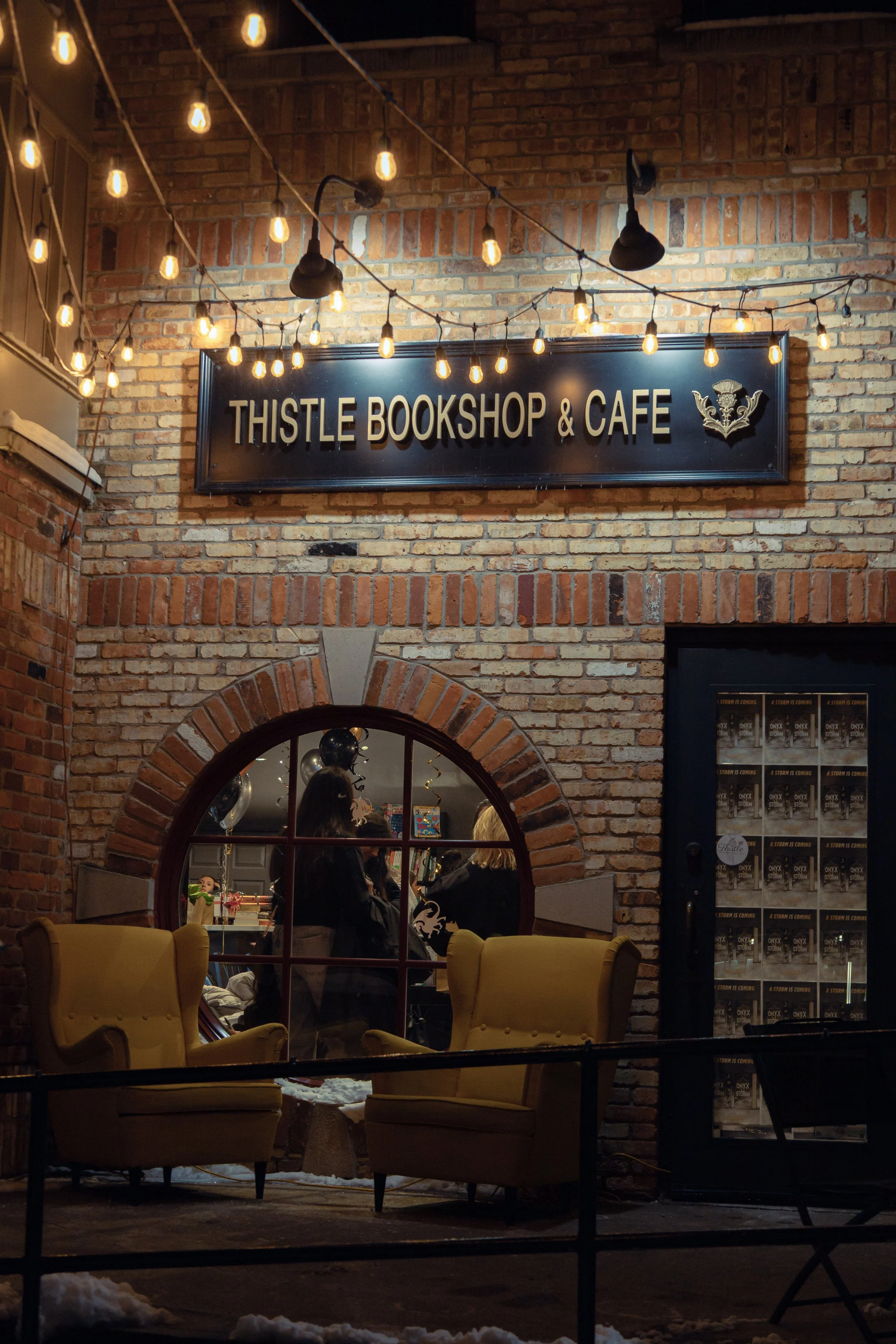 A brick storefront with a sign reading 'Thistle Bookshop & Café' and string lights hanging above. Through a window, people are visible inside the bookstore.
