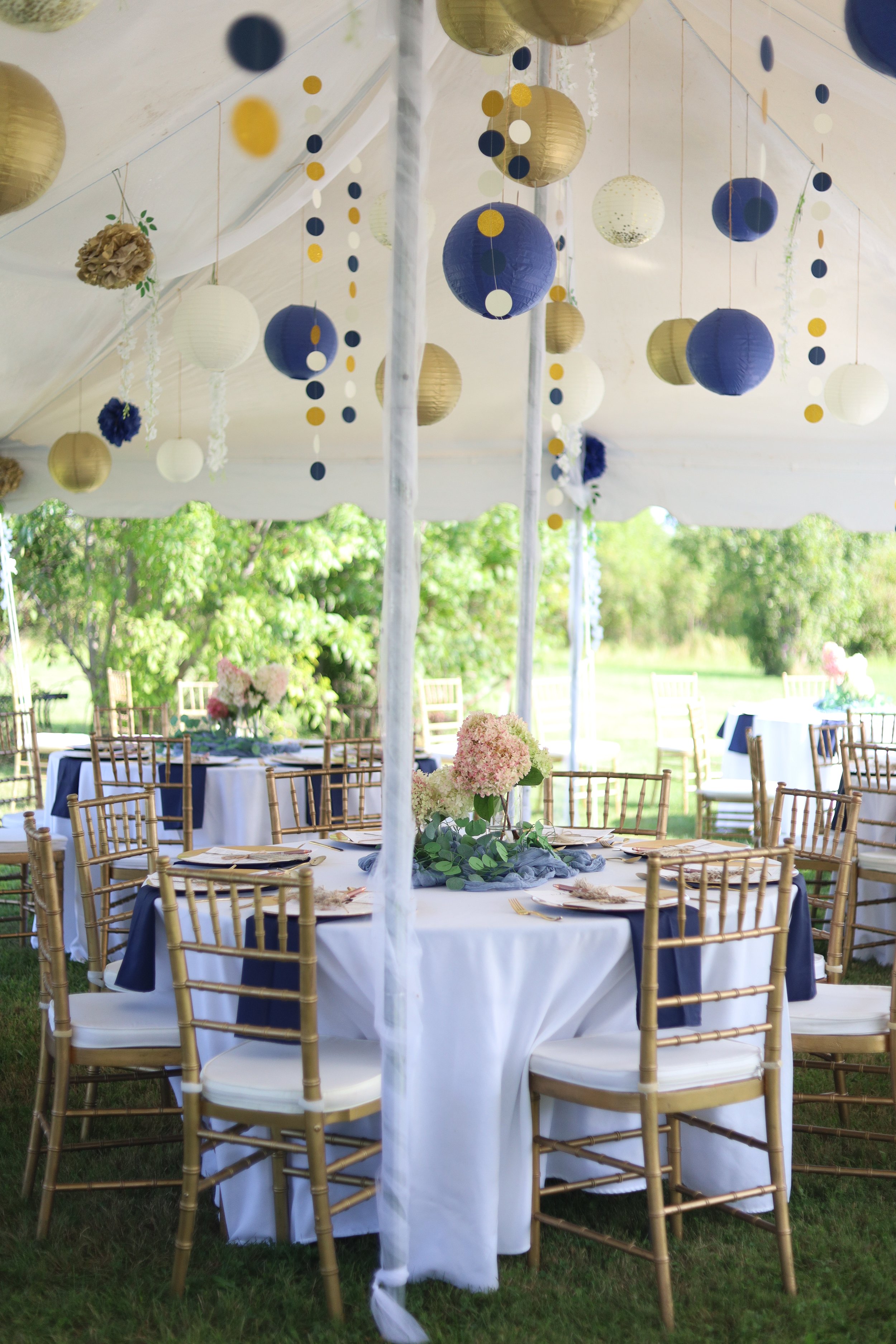 A decorated outdoor event tent with hanging paper lanterns in gold, white, navy blue, and dark blue, and gold and navy blue polka dot streamers. There are tables with white tablecloths, navy blue table runners, and centerpieces featuring pink and whi