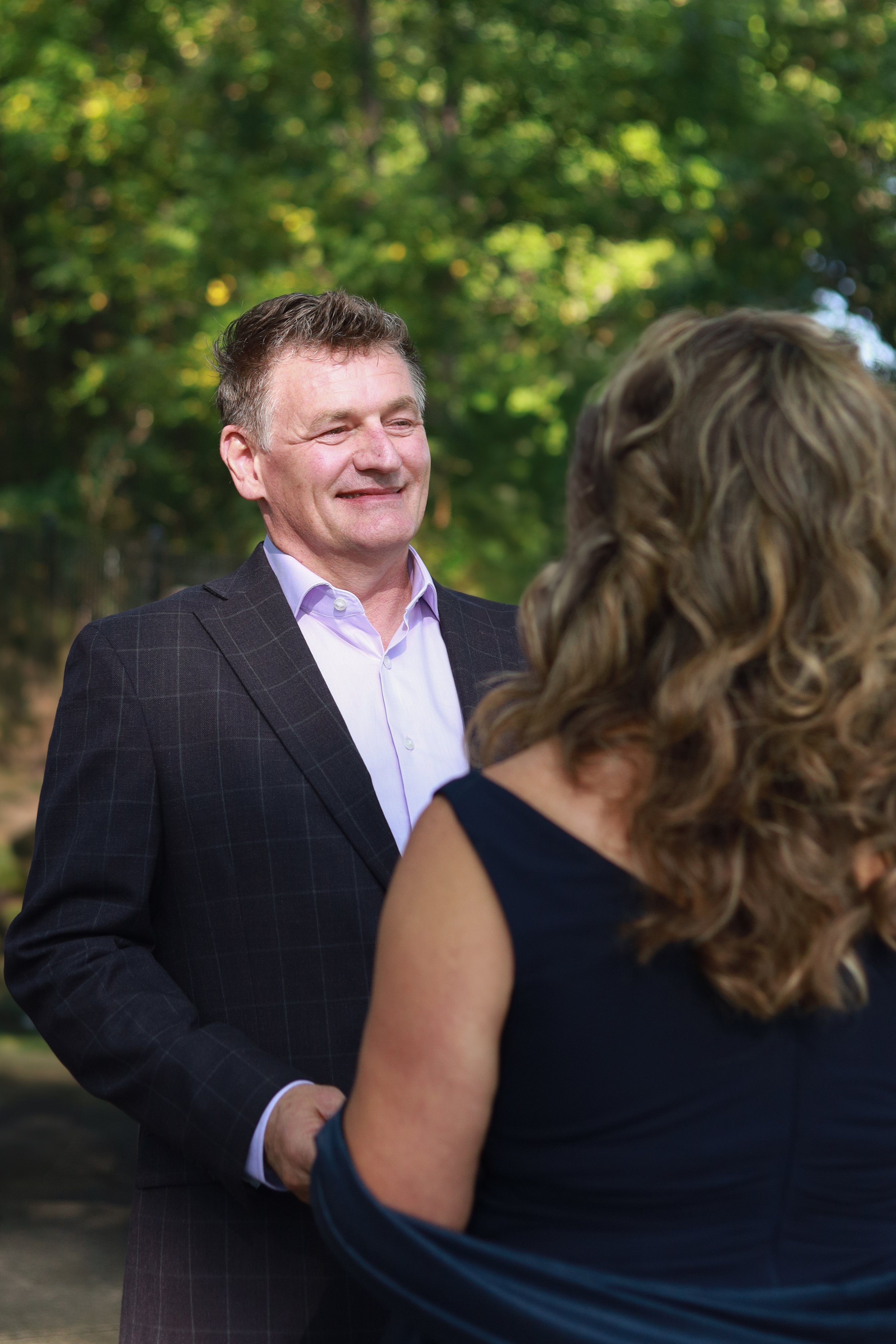 A man in a dark suit with a checkered pattern and a white shirt is smiling and talking to a woman with long curly blonde hair wearing a dark sleeveless dress, outdoors with green trees in the background.