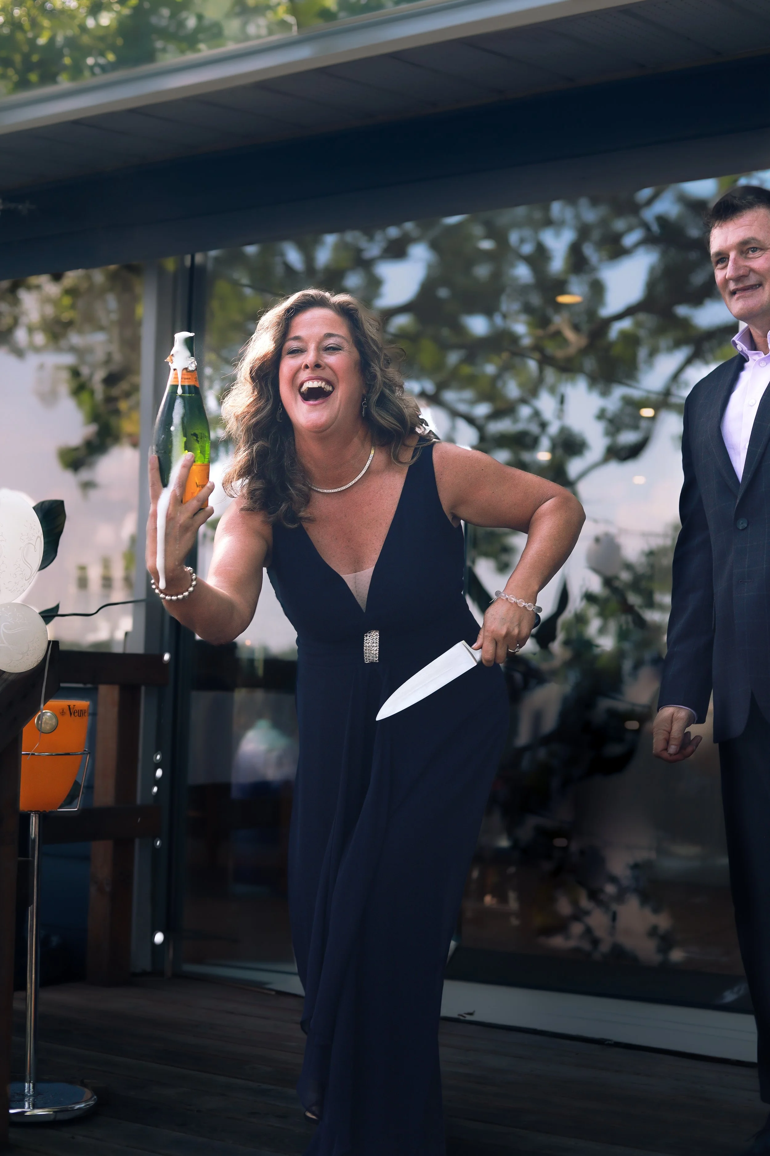 A woman in a navy blue dress laughing and holding a champagne bottle and a knife, standing outside near a glass door, with a man in a suit smiling beside her. They are at a celebration event.
