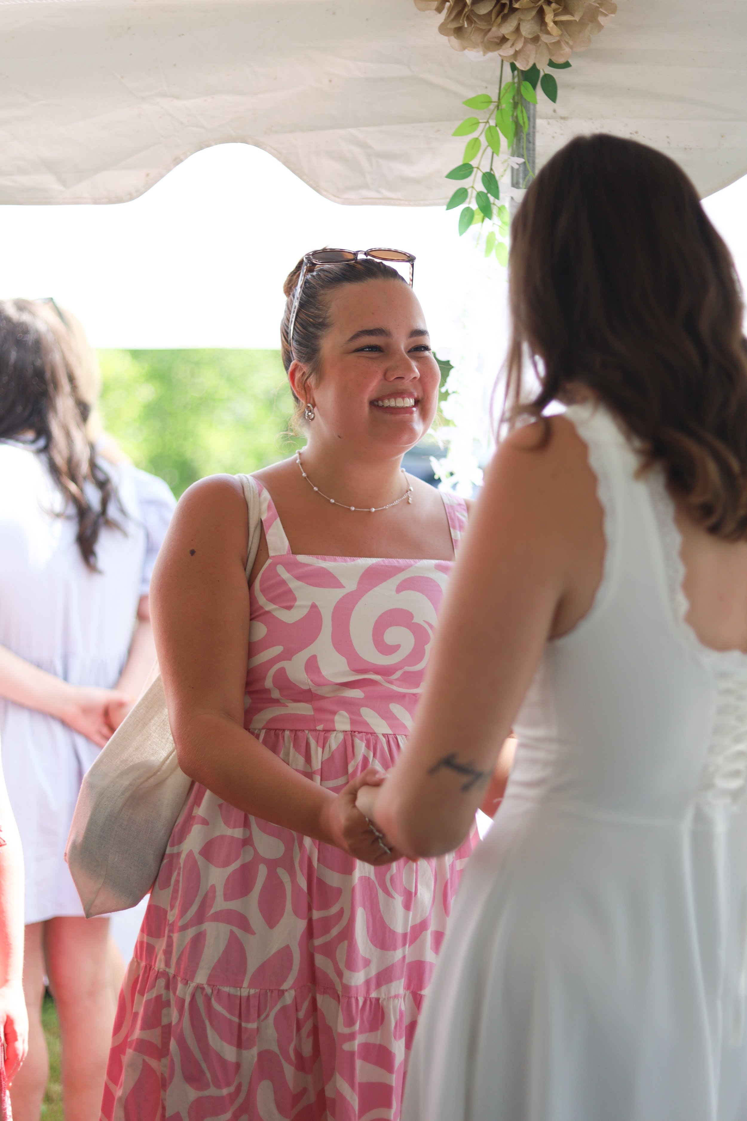 A woman in a pink and white patterned dress holding hands and smiling while talking to another woman in a white dress, with people in the background and greenery outside.