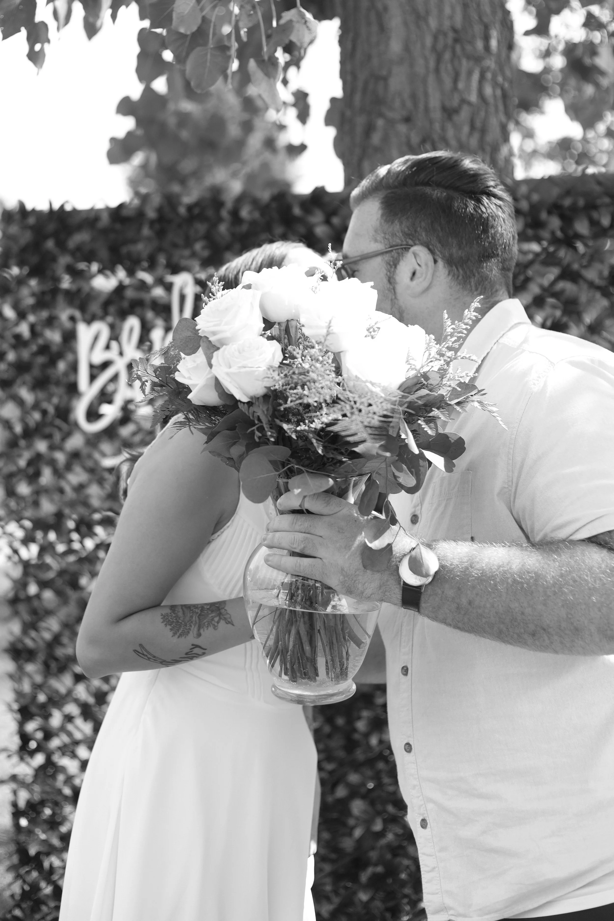 A black and white photo of a couple embracing outdoors, with the man holding a bouquet of flowers and the woman wearing a white dress. Their faces are close together, and they are in front of a hedge with a blurred background.