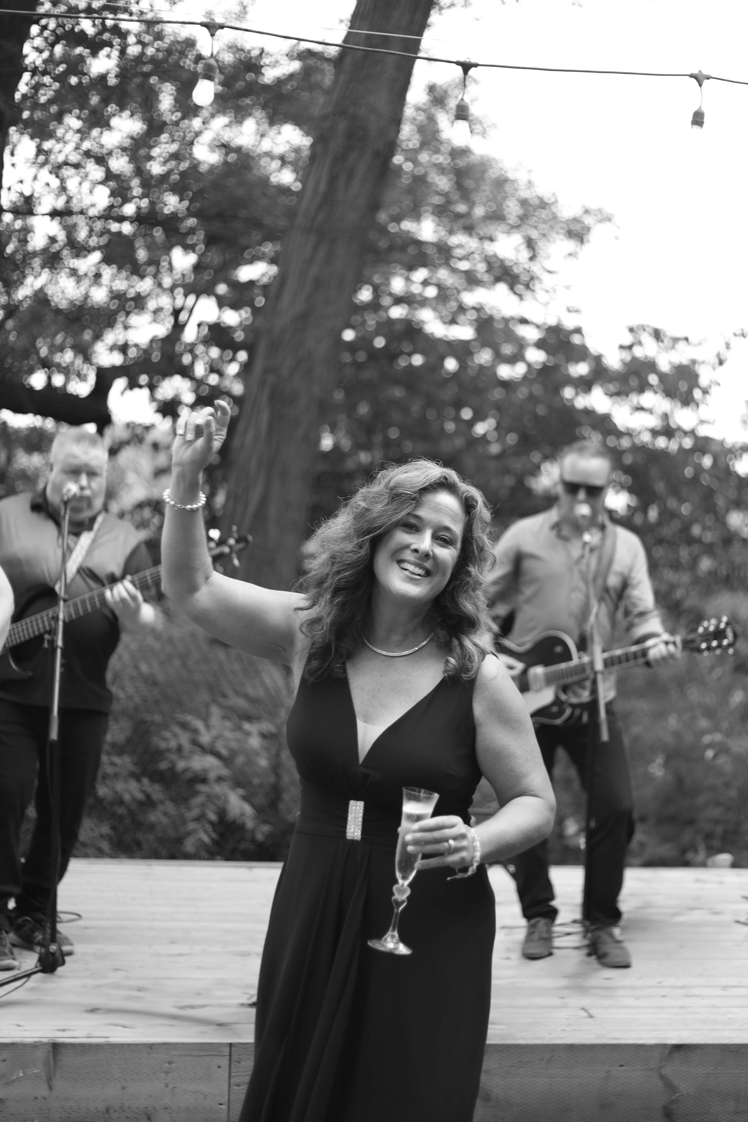 A woman in a black dress holding a glass of champagne, smiling and raising her hand, with two men playing guitars in the background, outdoors under string lights and trees.