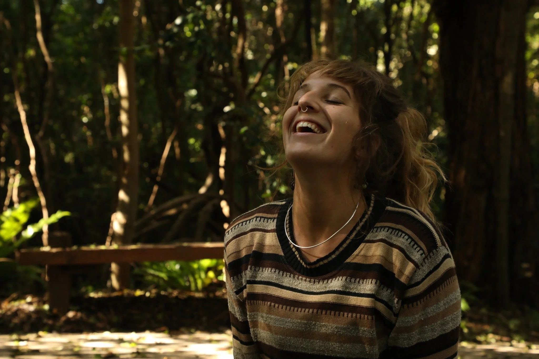A young woman with curly hair and a septum piercing smiling happily, sitting outdoors in a wooded area with tall trees and sunlight filtering through the leaves.