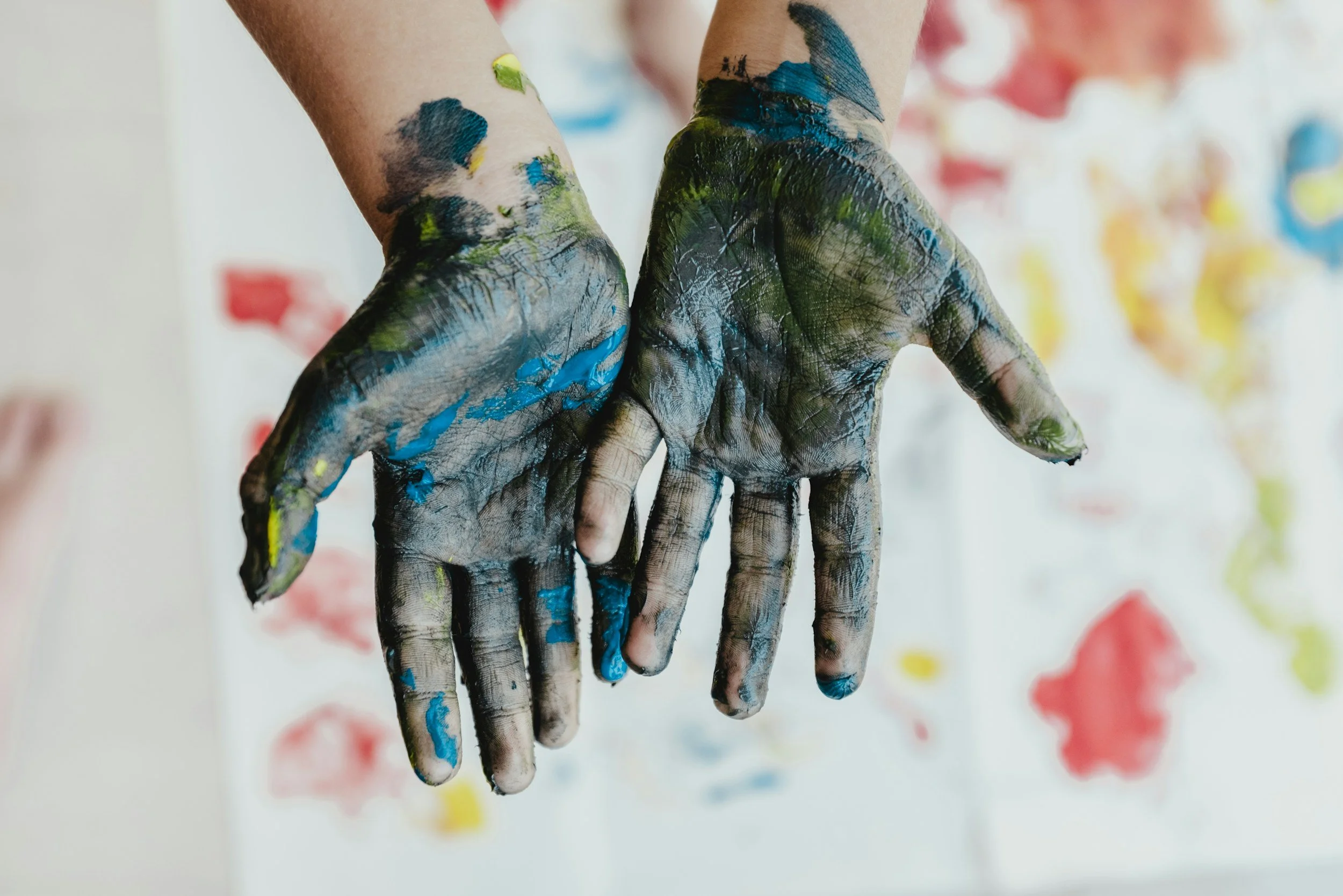 A person with hands covered in multicolored paint, sticking out in front of a colorful abstract background.