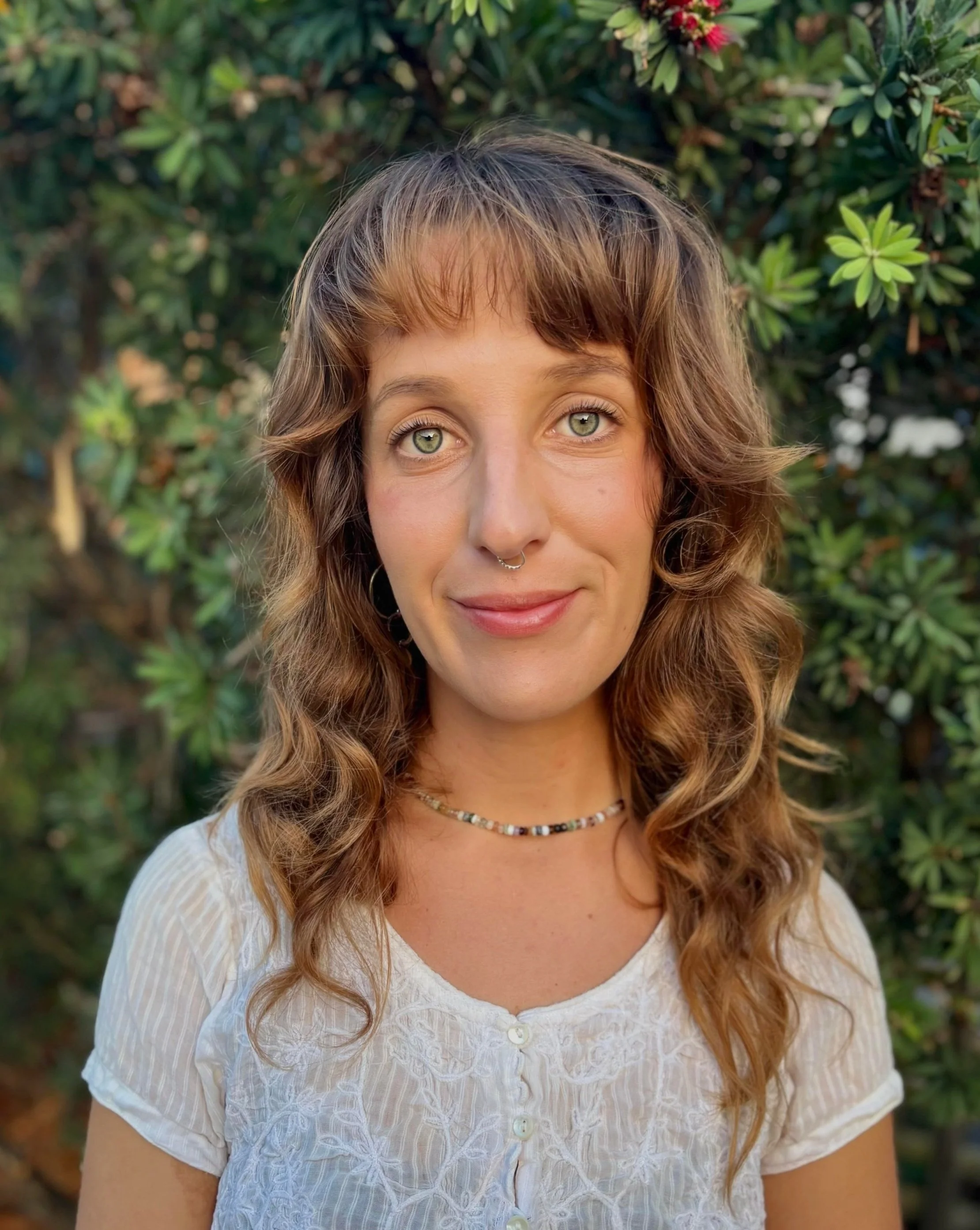 A young woman with light green eyes, wavy brown hair, and a septum piercing, wearing a white embroidered top and a colorful beaded necklace, standing outdoors in front of green foliage.