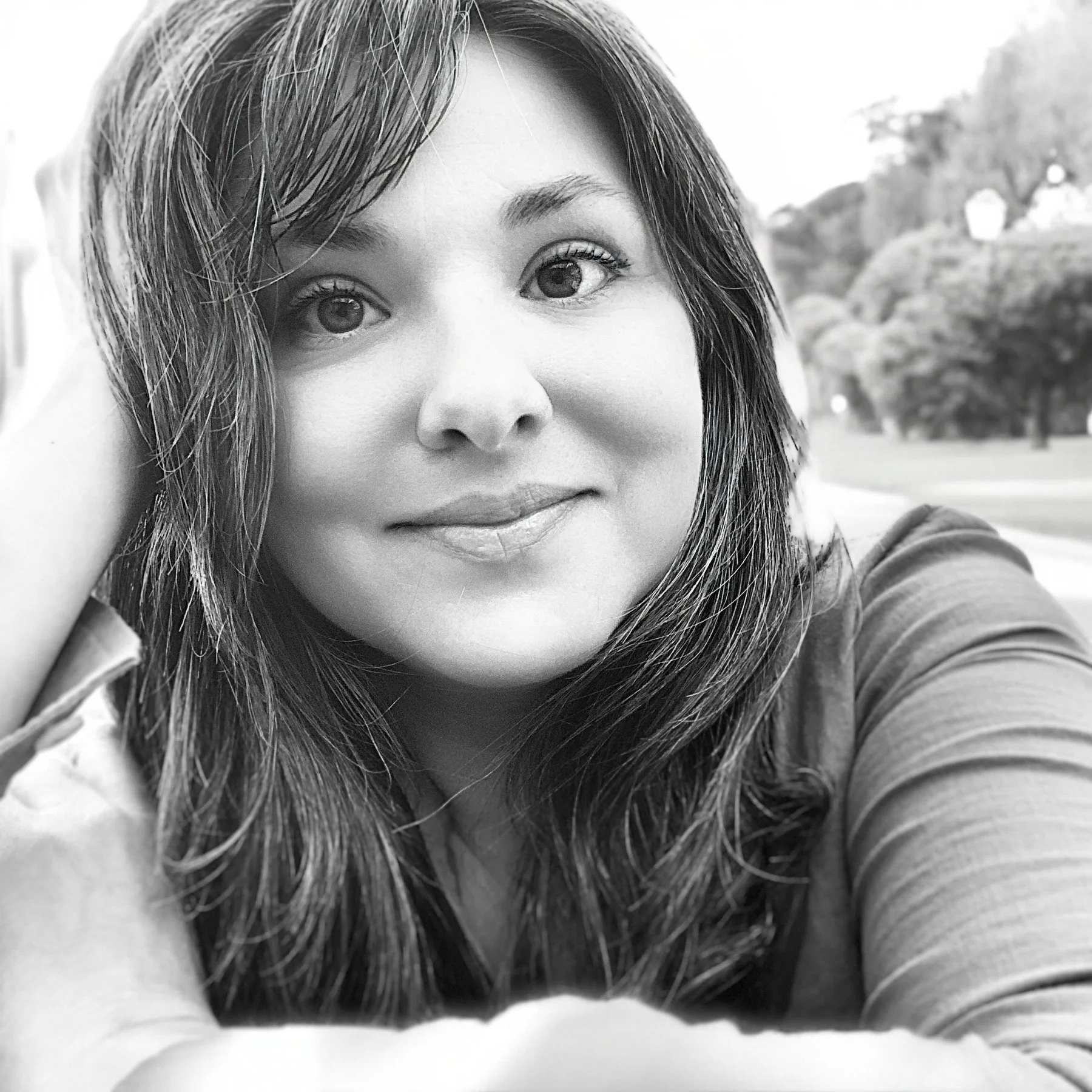 A black and white photo of a woman with shoulder-length hair, smiling softly, with her hand resting on her head, outdoors with trees in the background.