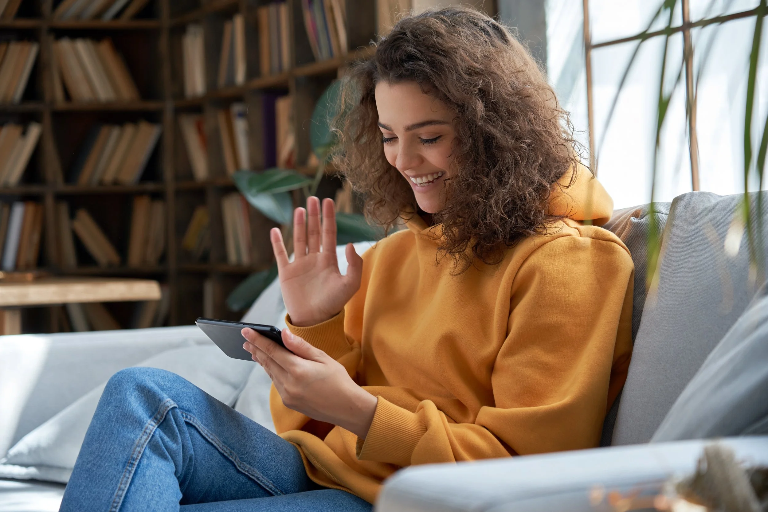 A young woman with curly hair in a yellow hoodie and jeans sitting on a couch, smiling and waving at her smartphone in a cozy, book-filled room.