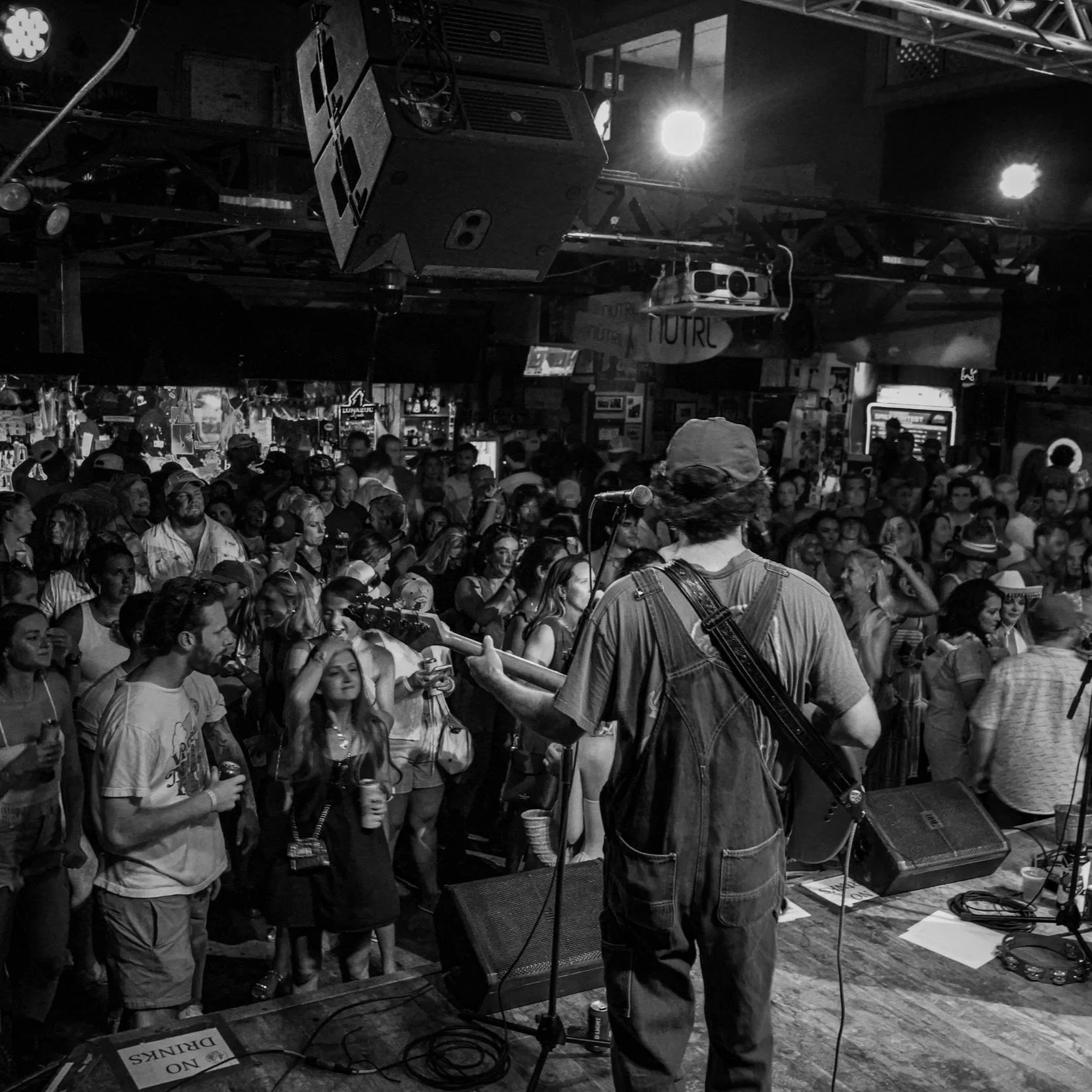 A black and white photo of a live music performance in a bar or club. The image shows a female singer on stage with a guitarist, with a large crowd watching. The stage is small with various lighting and sound equipment overhead.