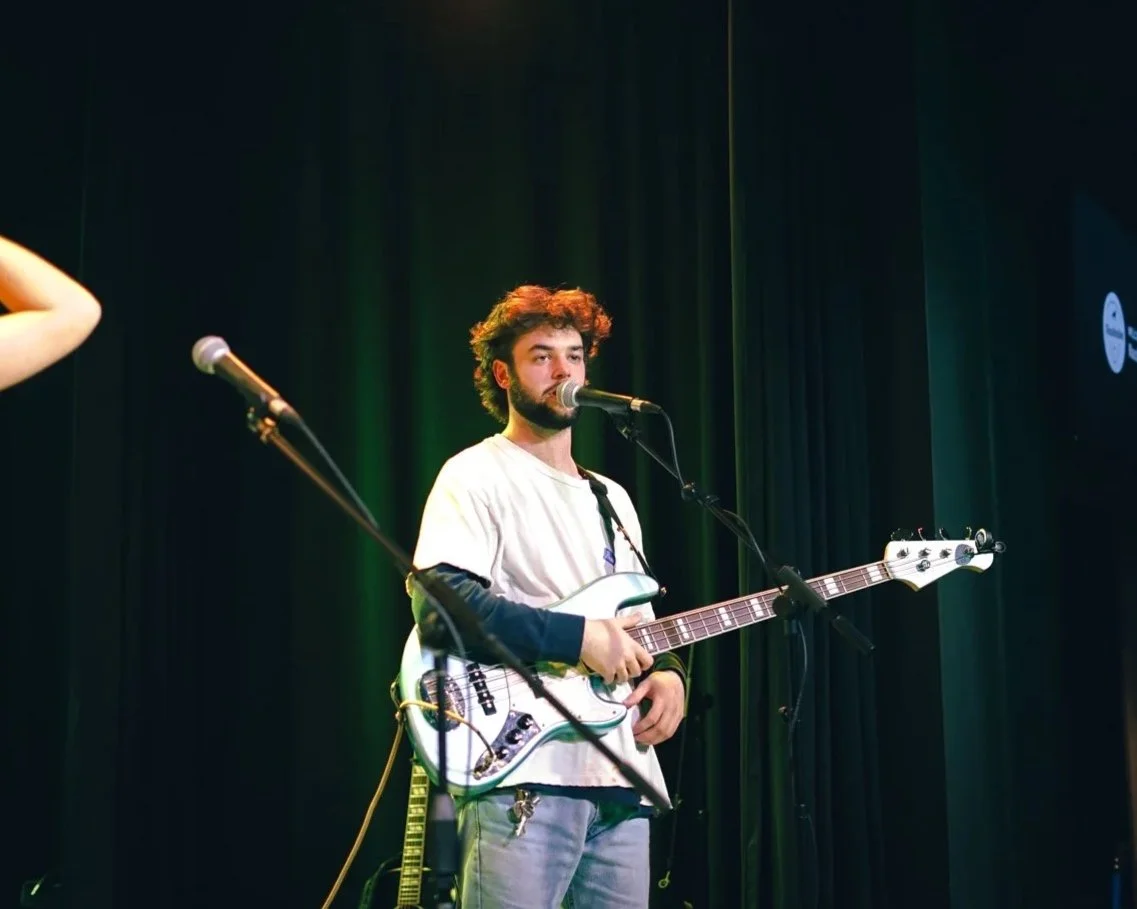 A male musician with curly hair and a beard, wearing a beige t-shirt and gray pants, standing on stage with a bass guitar, speaking into a microphone, with a female performer in a blue dress on the left adjusting her hair, stage lighting overhead, an