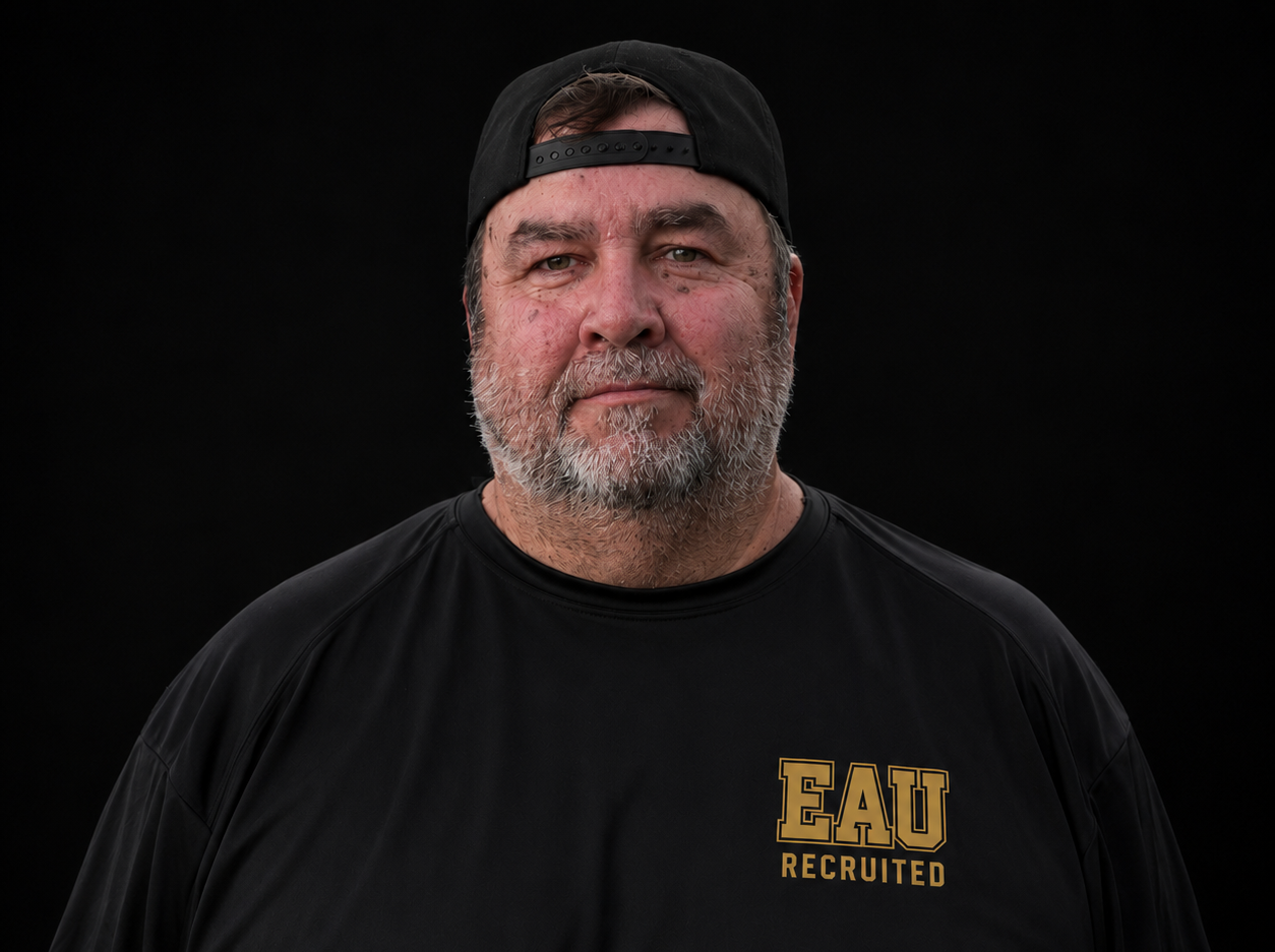 A middle-aged man with gray beard, wearing a black cap backwards and a black sports shirt with gold lettering that says 'EAU RECRUITED', standing against a black background.