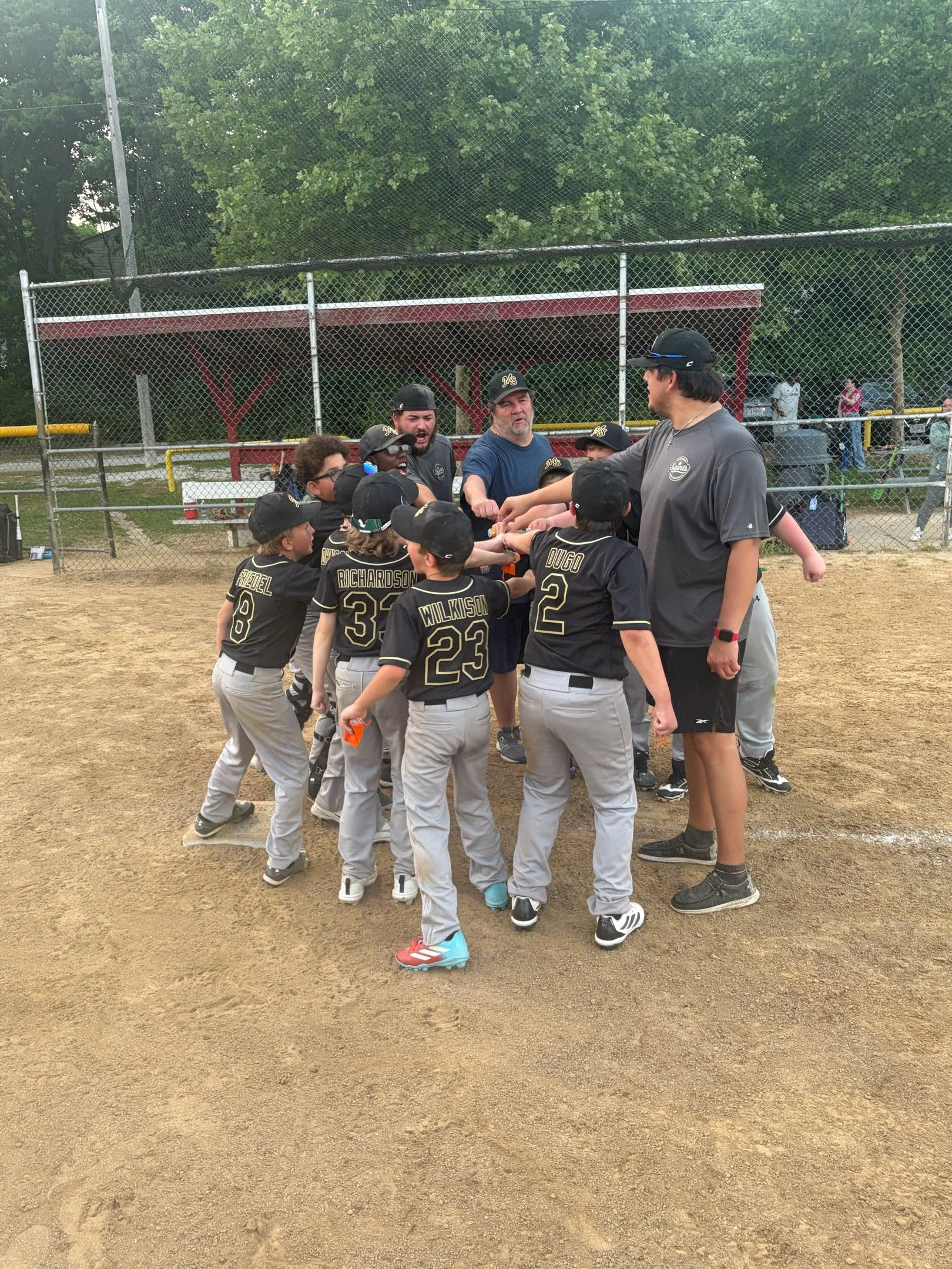 Youth baseball team gathering on the field with coaches after a game, players wearing black uniforms with gold lettering and numbers, some wearing caps, a chain-link fence in the background, and trees and fans in the distance.