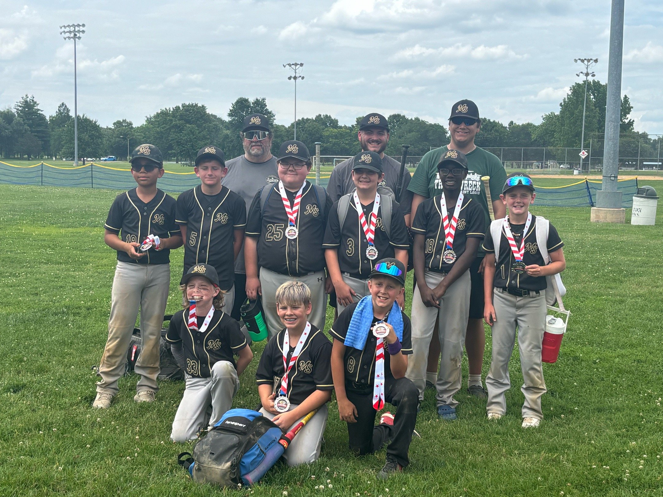 A group of children and coaches in baseball uniforms and caps standing and kneeling on a grassy field, smiling after a game, with medals around their necks and sports equipment. There are infield and outfield fences, trees, and light poles in the background.