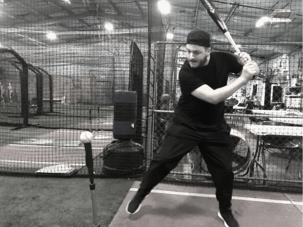 A man practicing baseball batting in an indoor batting cage, holding a bat and poised to hit a ball on a tee.